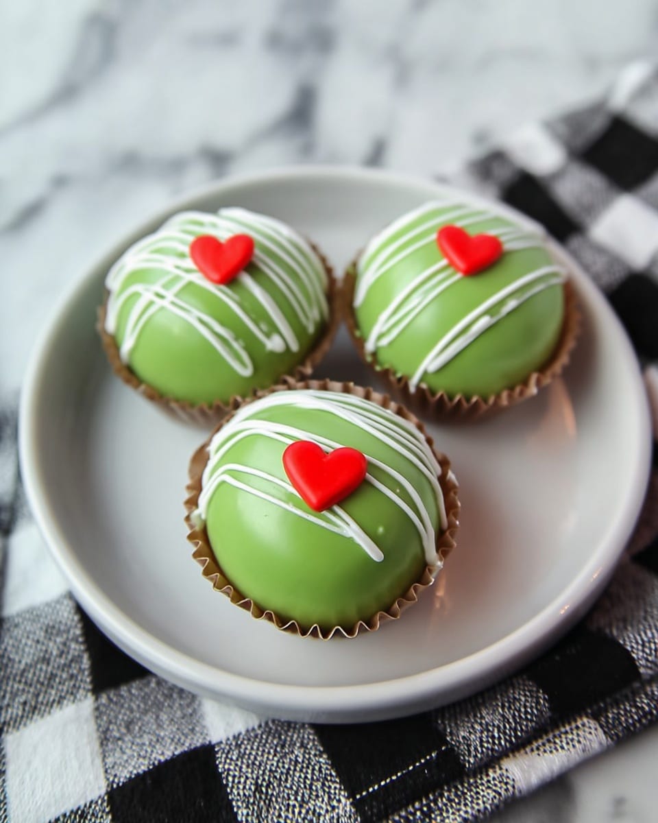 Three round desserts sit on a white plate, each one in a light brown paper cup. They have a smooth, shiny green shell with white thin drizzle lines going across the top. At the center of each dessert is a small, bright red heart-shaped decoration. The background shows a white marbled surface with a black and white checkered cloth partially visible underneath the plate. The overall look is clean and colorful, with the green and red standing out against the neutral background. photo taken with an iphone --ar 4:5 --v 7