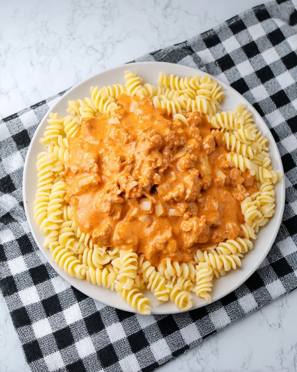 The image shows a white plate with two main layers. The bottom layer is a ring of pale yellow, spiral-shaped pasta arranged neatly around the plate’s edge. On top, there is a thick, creamy orange sauce with small pieces of meat and onions, spread in a mound in the center of the pasta, slightly spilling over onto the pasta edges. The plate is set on a white marbled surface with a black and white checkered cloth underneath part of it. Photo taken with an iphone --ar 4:5 --v 7