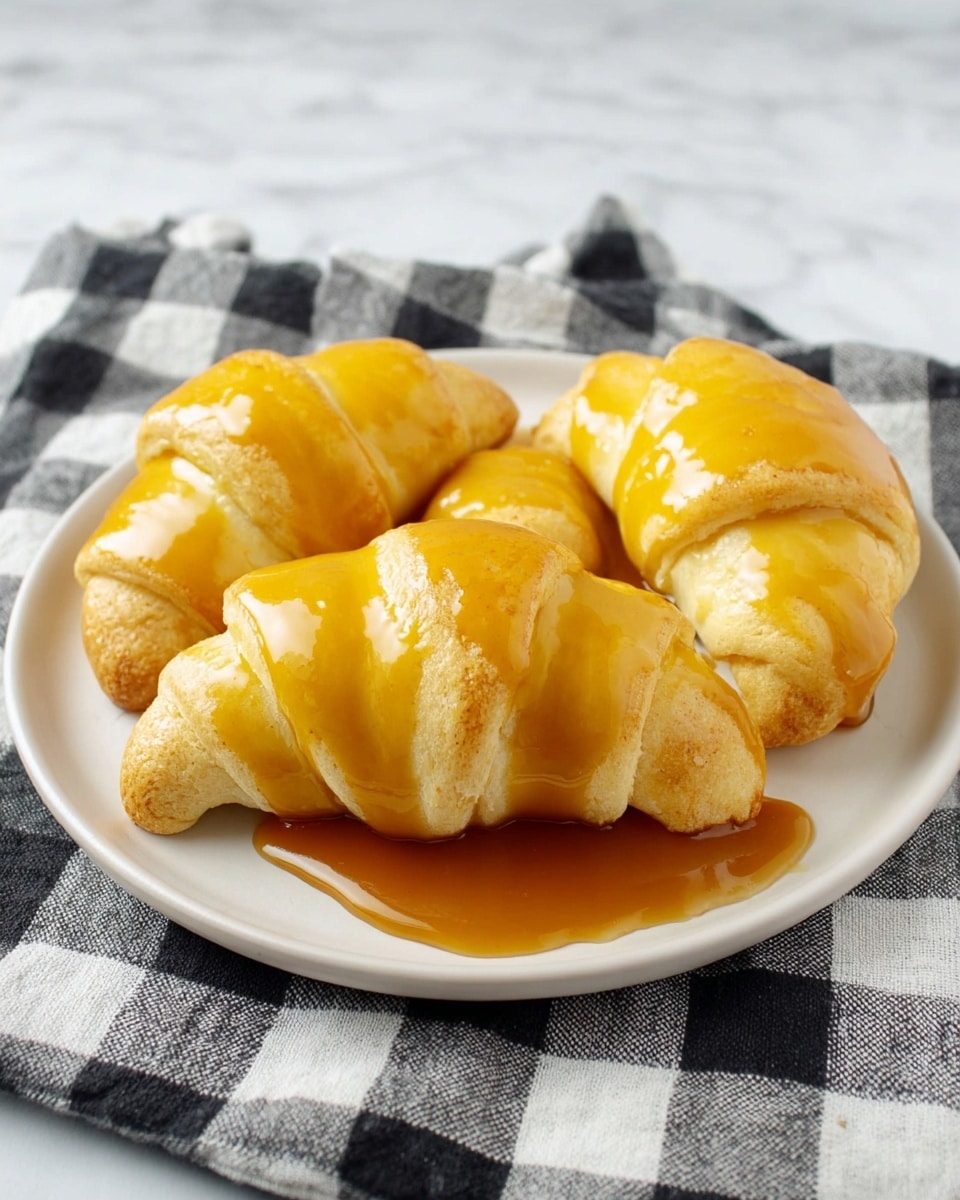 A white round plate holds four crescent rolls arranged close together, each roll is light golden with a soft texture, covered in thick, shiny amber caramel sauce that drips slightly down the sides and pools at the bottom of the plate; the plate rests on a black and white checkered cloth atop a white marbled surface. photo taken with an iphone --ar 4:5 --v 7