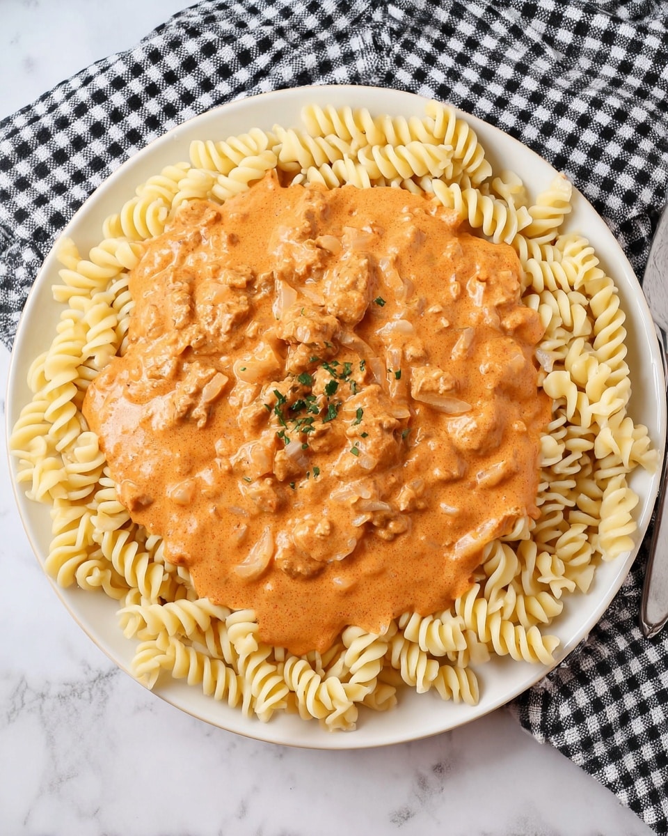 The image shows a white plate with two main layers of food. The bottom layer has yellow curly pasta arranged in a ring around the plate's edge, with some pasta pieces slightly overlapping. On top of the pasta, there is a thick orange creamy sauce with visible chunks of meat and small bits, covering the middle area of the pasta while some sauce spills around the edges onto the plate. The plate is placed on a black and white checked cloth, on a white marbled textured surface. photo taken with an iphone --ar 4:5 --v 7
