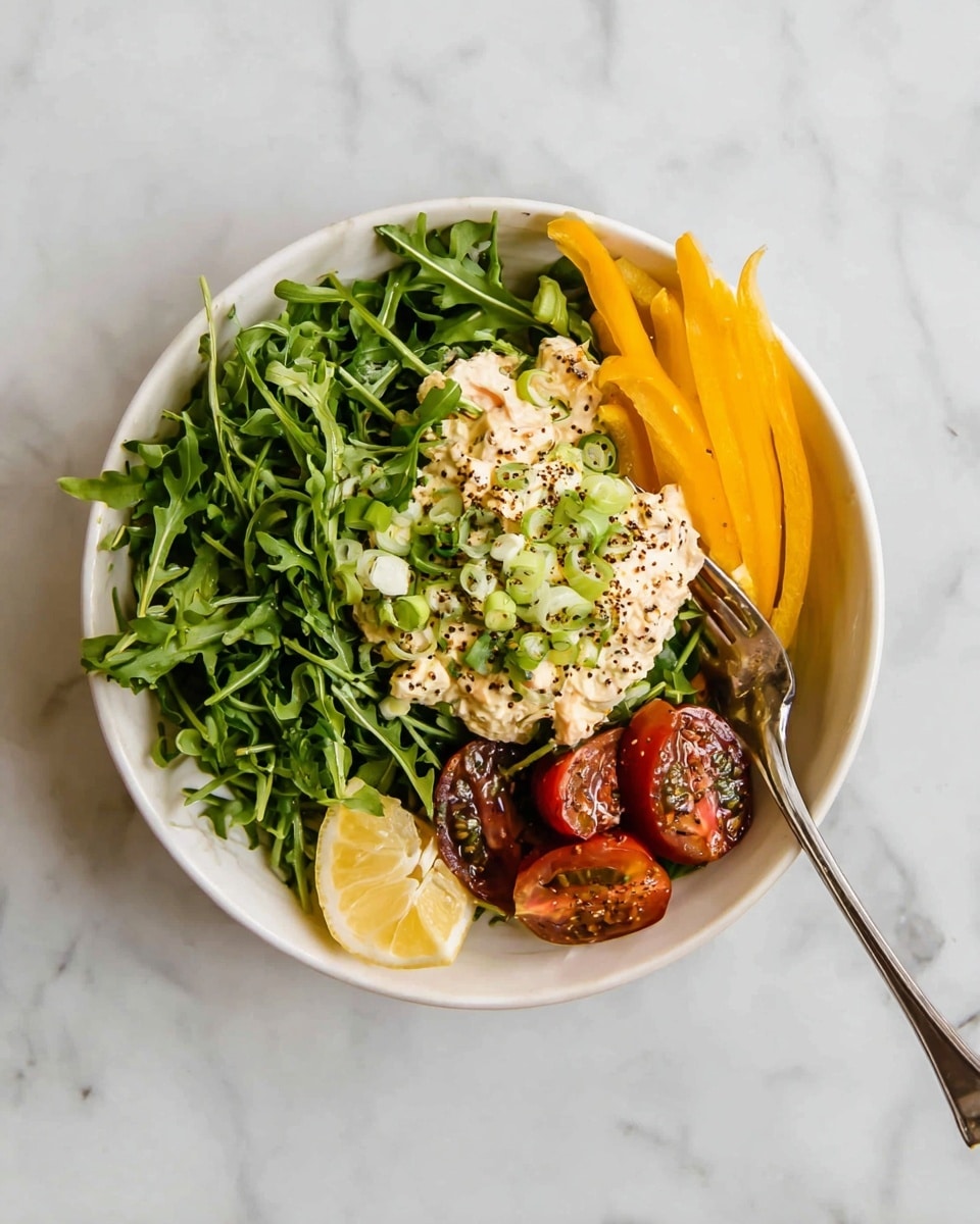 A white bowl filled with a base layer of fresh, green arugula leaves covering the entire bottom. On the right side, a small pile of sliced yellow bell pepper sticks stands upright. In the center, there is a creamy, light beige scoop of a mixed salad topped with thinly sliced green onions and sprinkled with seasoning. To the bottom right of the creamy salad, there are halved dark red grape tomatoes, also sprinkled with seasoning. A small wedge of lemon rests at the edge near the tomatoes. A silver fork is placed inside the bowl on the right side, leaning against the edge. The bowl sits on a white marbled surface. photo taken with an iphone --ar 4:5 --v 7