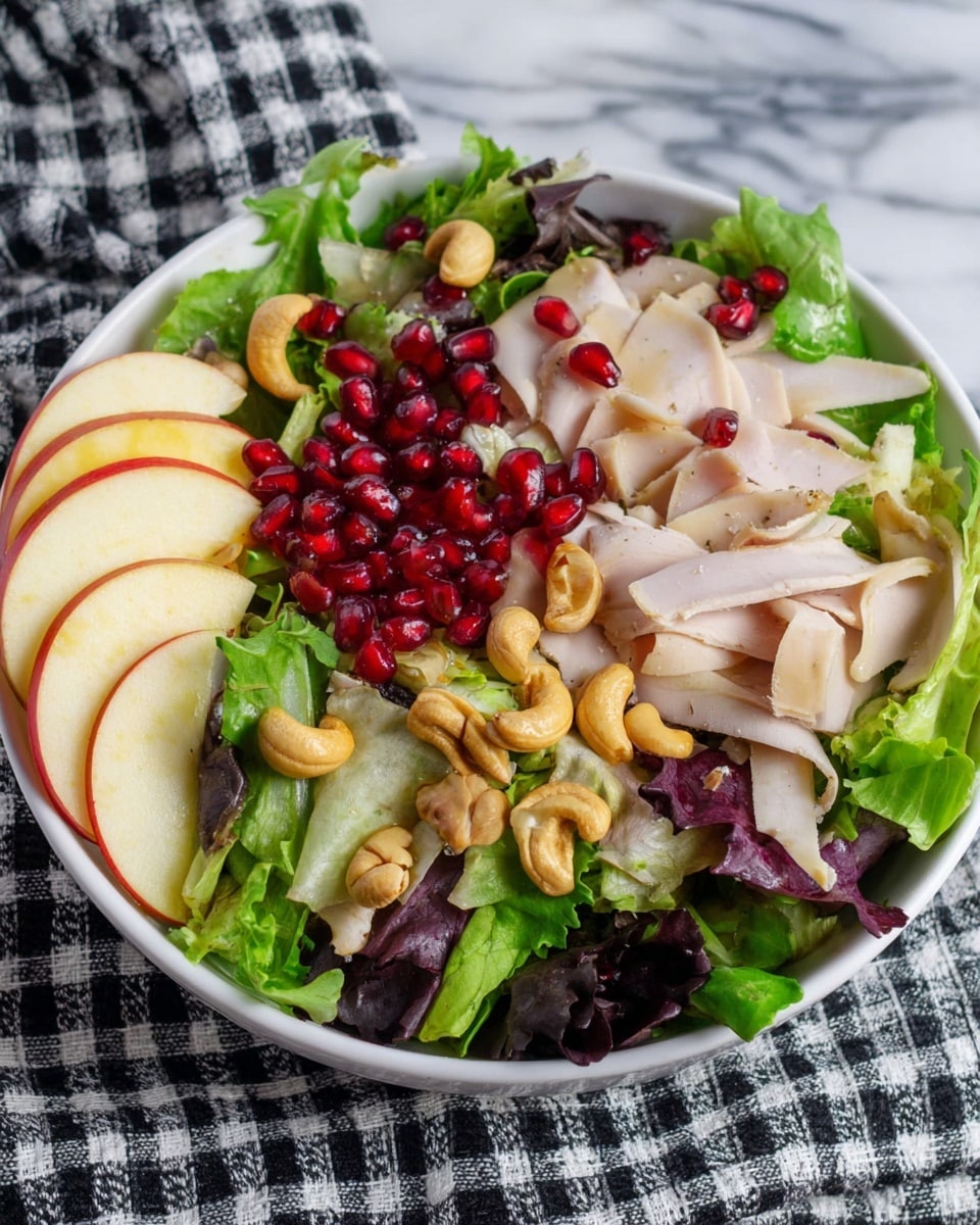A white bowl holds a fresh salad with multiple layers starting with a base of mixed green leaves in different shades of green and some dark purple. On top of the greens are light beige thin slices of turkey, scattered bright red pomegranate seeds, and tan cashew nuts placed mainly to one side. Thin slices of pale yellow-green apple with red edges are arranged along the left side of the bowl. The bowl sits on a white marbled surface with a black and white checked cloth partially under the bowl. photo taken with an iphone --ar 4:5 --v 7