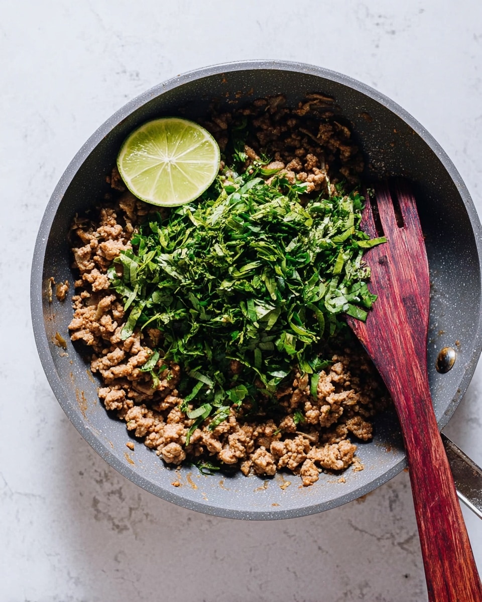 A grey frying pan filled with cooked ground meat showing a light brown and slightly greasy texture, topped with a generous pile of chopped fresh green herbs in the center, and a half-cut light green lime resting on top of the herbs. A wooden spatula with a deep reddish-brown color is placed inside the pan, partially covering the herbs and meat. The pan is set on a white marbled textured surface. photo taken with an iphone --ar 4:5 --v 7