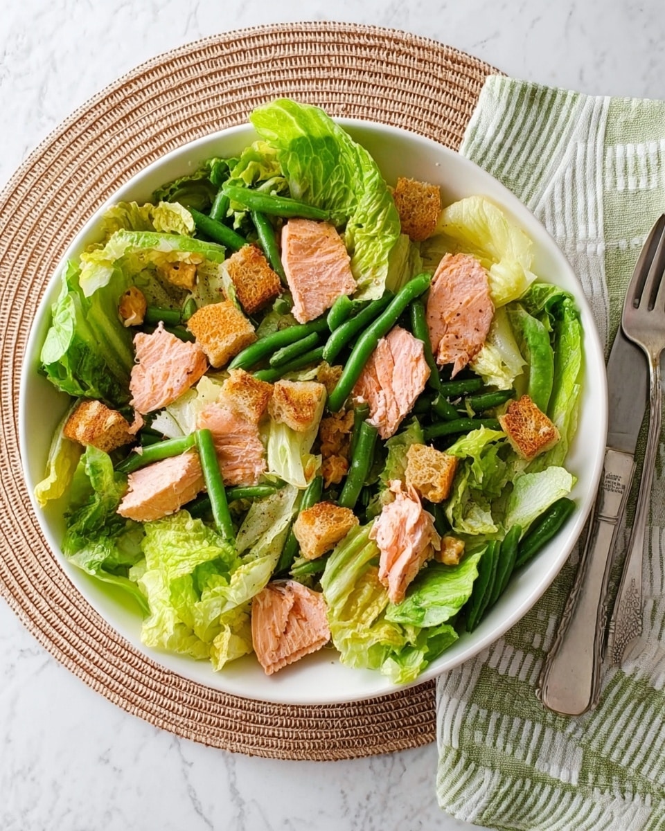 A white round plate holds a fresh salad with several layers: the bottom layer is green romaine lettuce with large, crisp leaves covering the plate, topped with pieces of cooked pink salmon scattered evenly. On top, there are long, thin green beans placed in a slightly haphazard way, adding texture and color contrast. Small, golden-brown croutons are sprinkled across the salad, adding a crunchy detail. The whole plate is set on a white marbled surface with a round woven mat underneath, and to the left of the plate, a stacked set of cutlery wrapped in a green-striped napkin is visible. photo taken with an iphone --ar 4:5 --v 7