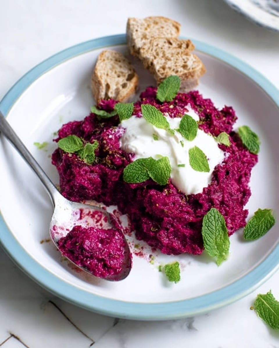 The image shows a white plate with a thin light blue rim, placed on a white marbled surface. On the plate, there is a single thick layer of bright red beetroot spread with a rough texture, topped unevenly by a white creamy sauce with a smooth texture. Small green mint leaves are scattered on top of both the beet spread and sauce, adding a pop of color. There are two pieces of torn light brown bread on the right side of the plate. A spoon with some of the beet spread and white sauce is resting on the plate with a woman's hand holding it. Photo taken with an iphone --ar 4:5 --v 7