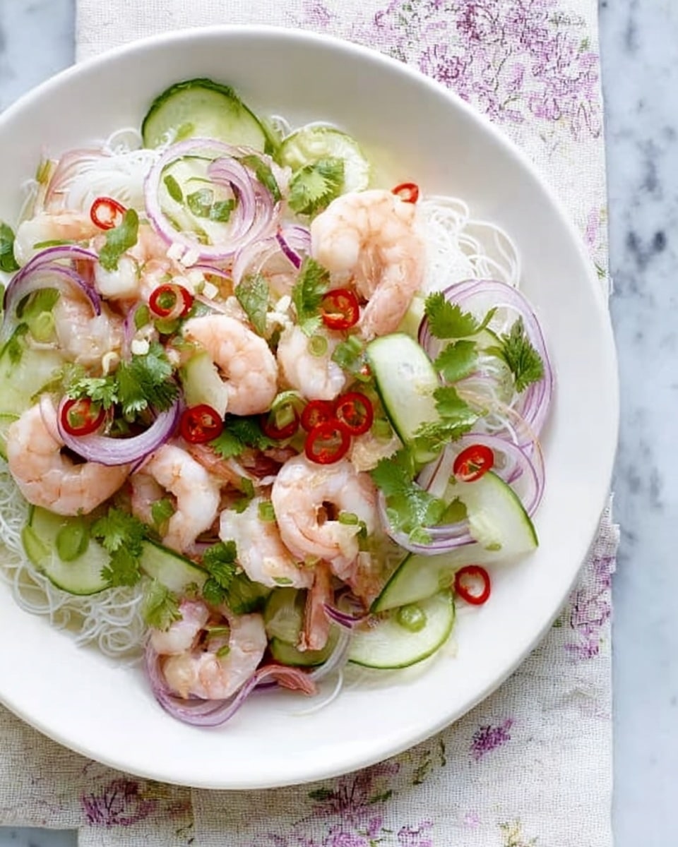 A white round plate holds a fresh shrimp salad with several layers; the bottom layer is thin white rice noodles, topped with light pink shrimp arranged loosely, thinly sliced red onions adding purple color, and thin green cucumber ribbons curled gently on top. Small red chili slices, green cilantro leaves, and tiny green mint leaves are scattered throughout, creating a colorful and fresh look. The plate is placed on a table covered with a muted light fabric, over a white marbled surface. Photo taken with an iphone --ar 4:5 --v 7