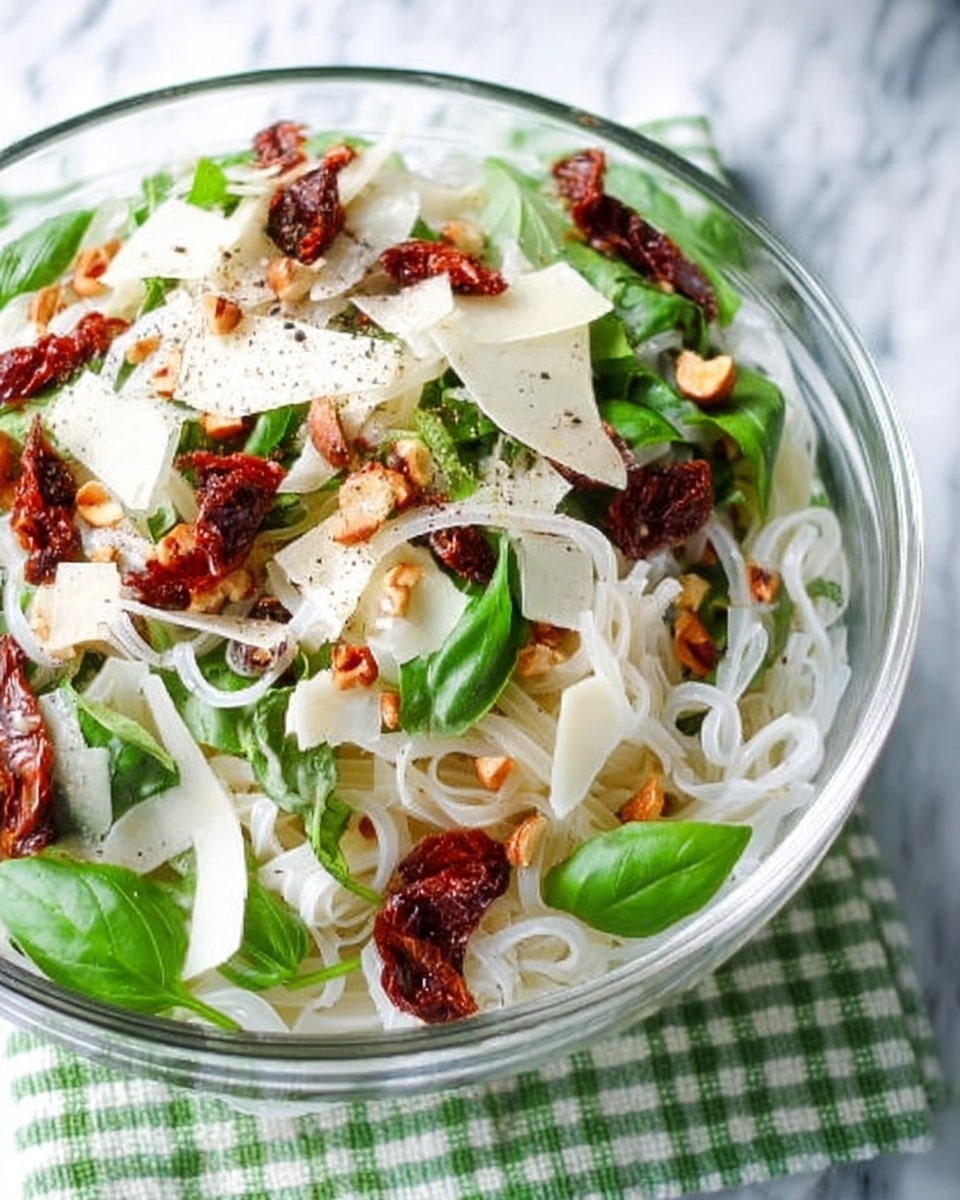 The image shows a clear white bowl filled with a layered salad on a white marbled surface with a green and white checkered cloth beneath. At the bottom are thin white noodles forming the first layer, soft and tangled. On top of this lies a mix of fresh green basil leaves and scattered pieces of dried sun-dried tomatoes that are deep red and wrinkled. Thin white slices of cheese are spread around the salad, with a few pale round nuts sprinkled throughout. The whole dish is lightly sprinkled with black pepper, adding tiny dark specks over the top photo taken with an iphone --ar 4:5 --v 7