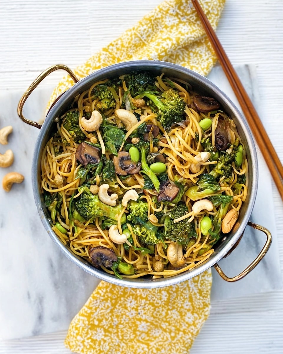 A metal pan filled with a layer of cooked yellow noodles at the bottom, topped with a mix of green broccoli florets, dark green leafy vegetables, sliced brown mushrooms, white cashew nuts, and small round green peas spread evenly across. The pan has a wooden handle at the top. The setting includes a white marbled surface with wooden chopsticks lying next to the pan and a white and yellow patterned cloth in the background. photo taken with an iphone --ar 4:5 --v 7