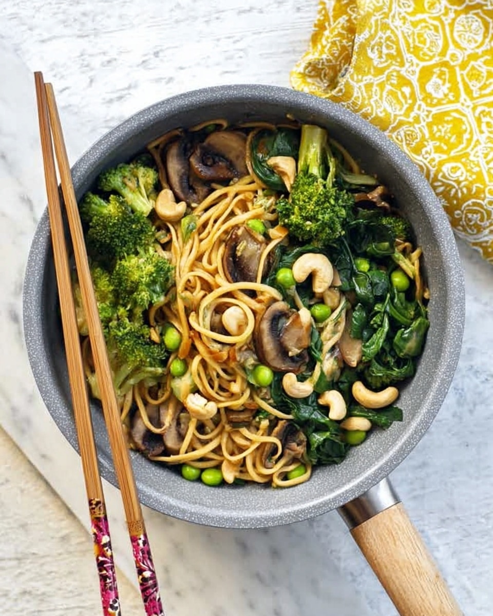 A close-up of a round metal pan filled with stir-fried noodles. The noodles are light brown and tangled, forming the base layer. On top, there are bright green broccoli florets and dark green leafy vegetables scattered evenly. Light brown mushroom slices with a soft texture are mixed in, along with white cashew nuts sprinkled all over. Small green peas add extra color and texture. The pan rests on a white marbled surface with a pair of wooden chopsticks on the right side and a yellow cloth with a green pattern in the upper left corner. Photo taken with an iphone --ar 4:5 --v 7