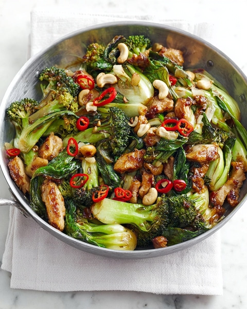 A round silver bowl filled with a colorful stir-fry dish sits on a white napkin over a white marbled surface. The dish has three main layers: the bottom layer is mixed green vegetables like broccoli and bok choy with some light brown cooked pieces of tofu or chicken, the middle layer includes bright red thin chili strips and sliced green onions adding texture, and the top layer is scattered with pale beige cashew nuts and a few yellow beans. The vegetables look crisp and slightly charred, with a glossy finish from the sauce. photo taken with an iphone --ar 4:5 --v 7