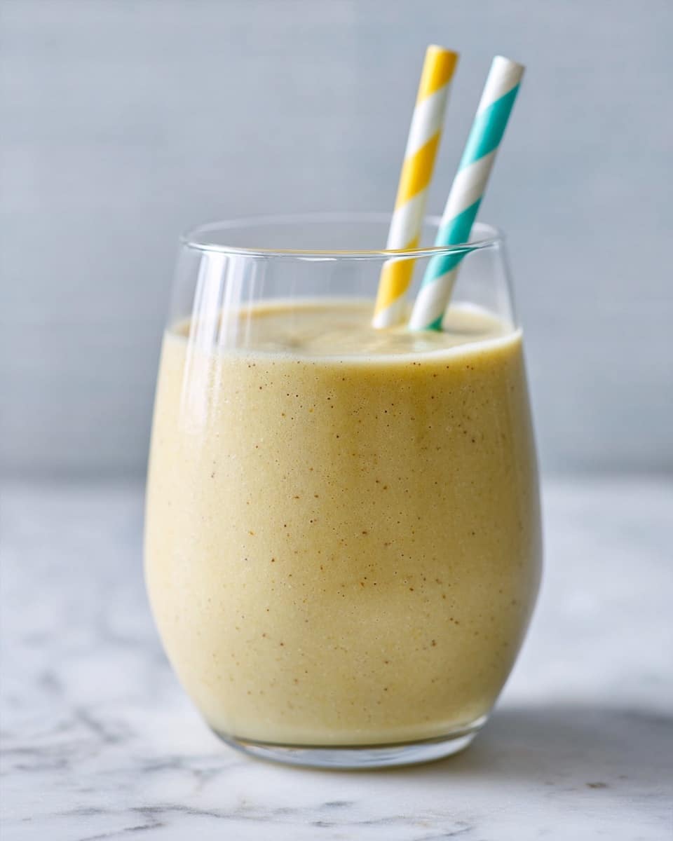 A clear glass filled with a creamy, light yellow smoothie sits centered on a white marbled surface. The smoothie has a smooth texture and fills most of the glass, leaving a small gap at the top. Two straws, one blue with white stripes and one yellow with white stripes, are placed inside the smoothie, leaning slightly to the right. The background is softly blurred, highlighting the glass and smoothie. Photo taken with an iphone --ar 4:5 --v 7
