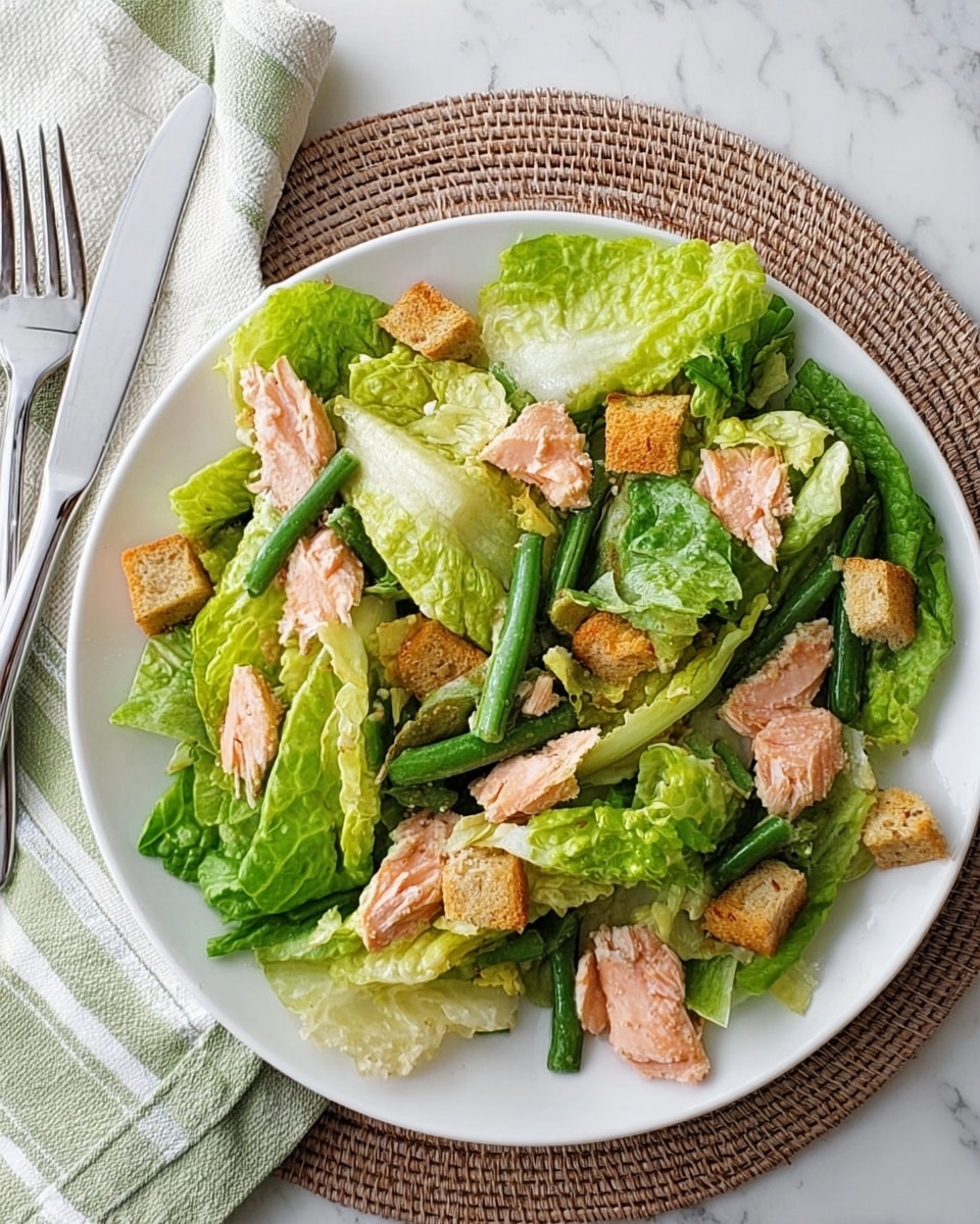 A white plate on a white marbled surface holds a salad with several layers: large green romaine lettuce leaves form the base layer, topped unevenly with light pink chunks of cooked salmon, long thin green beans, and small golden-brown croutons. The salad looks fresh with a simple arrangement without dressing visible. The plate is set on a round woven placemat. Photo taken with an iphone --ar 4:5 --v 7