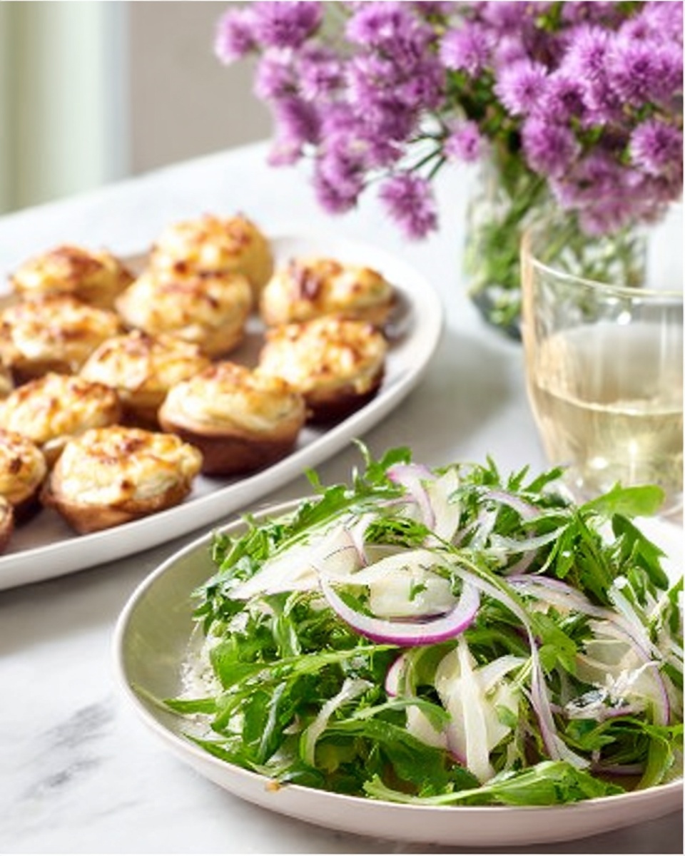 The image shows a fresh salad placed in a white bowl on a white marbled surface, with layers of bright green leafy arugula, thinly sliced fennel, and thin red onion rings mixed throughout, giving a mix of green, white, and purple colors. Behind the salad, there is a white plate stacked with small round bread rolls topped with a golden, baked cheese layer, each roll having a crispy, browned texture on top. To the right, there is a glass with a light-colored drink, and in the background, a vase with light purple flowers adds a soft touch to the scene. Photo taken with an iphone --ar 4:5 --v 7