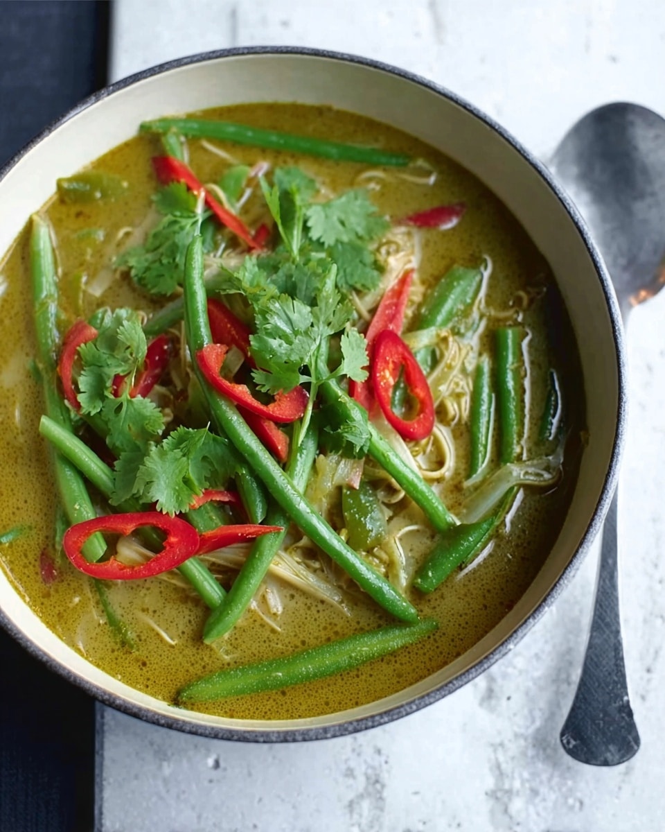 A deep round white bowl on a white marbled surface holds a light green curry soup. Inside the soup, there are multiple layers of bright green beans and thin red chili slices scattered on top. Visible also are cilantro leaves spread across the curry, adding a fresh green color contrast. The soup has a smooth texture with some noodles slightly visible beneath the green beans. To the right of the bowl, there is a large spoon resting on the surface. photo taken with an iphone --ar 4:5 --v 7