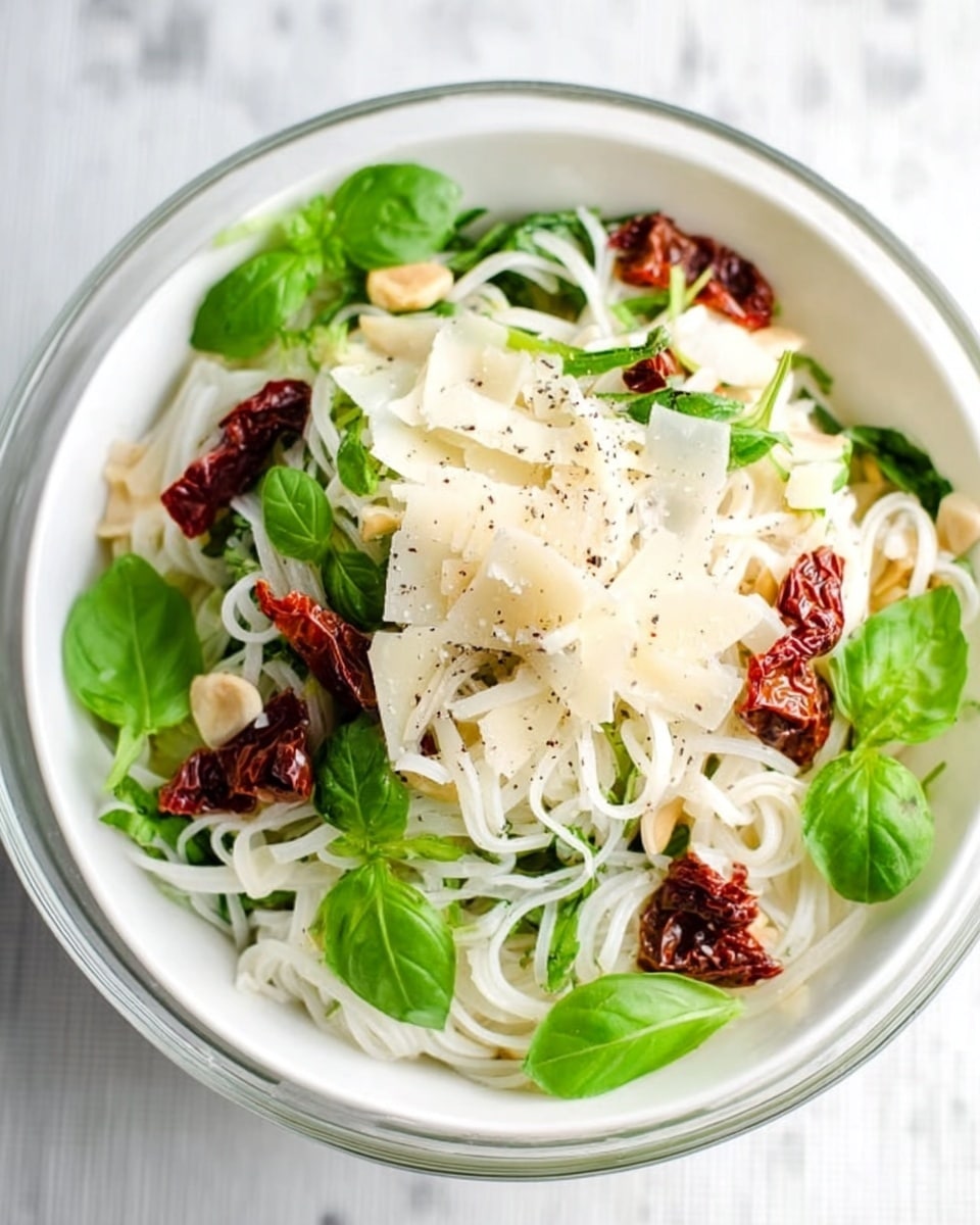 A clear white bowl filled with a fresh pasta dish, showing two main layers: the bottom layer of soft, white pasta ribbons mixed lightly with thin white cheese slices, and the top layer of green basil leaves with scattered dark red sun-dried tomato pieces and tiny beige nuts. The dish is sprinkled with black pepper bits, all placed on a white marbled surface with a green and white checkered cloth partially visible under one side. photo taken with an iphone --ar 4:5 --v 7