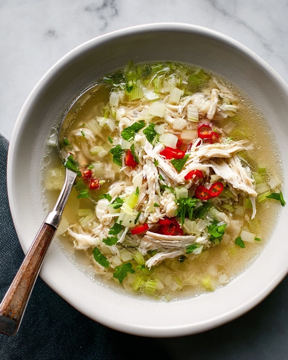 A white bowl filled with clear chicken soup showing three main layers: at the base, translucent onion slices and small light green pieces; in the middle, shredded white chicken and thin slices of red chili peppers; on top, a small heap of cooked brown rice and chopped fresh green herbs. A silver spoon with a wooden handle rests inside the bowl, leaning against the side. The bowl sits on a white marbled surface. photo taken with an iphone --ar 4:5 --v 7