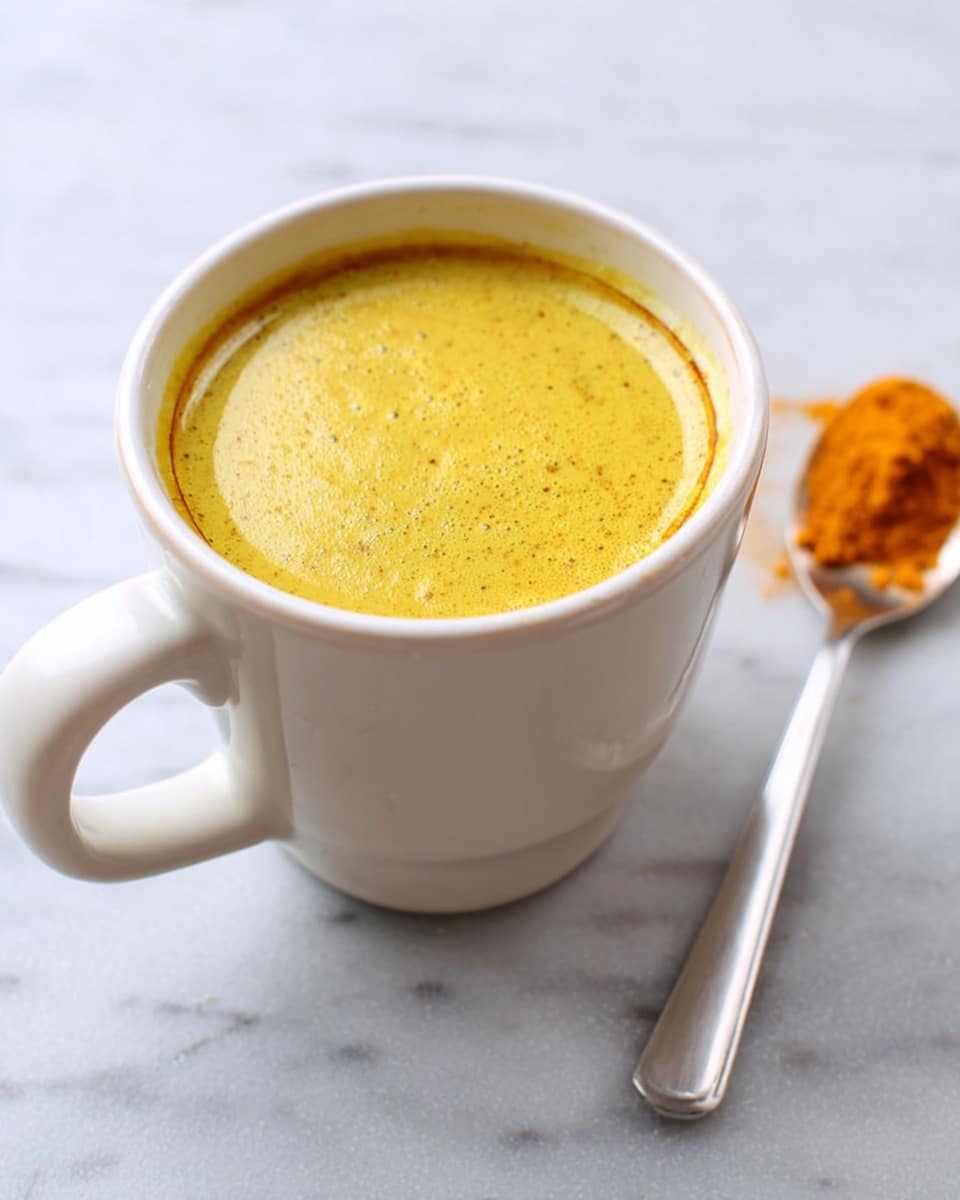A white mug filled with a smooth, frothy, golden-yellow liquid sits on a white marbled surface. The surface of the drink has a few small bubbles scattered across it, creating a slightly textured look. Behind the mug, a silver spoon rests flat on the surface with a small pile of bright orange powder on its bowl, adding a contrasting color to the scene. The overall image has a clean and simple look. photo taken with an iphone --ar 4:5 --v 7