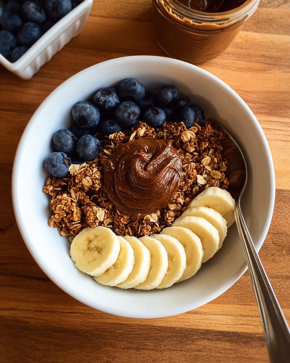 The image shows a white bowl filled with granola on the top half, fresh blueberries in the middle, and a dollop of chocolate almond butter sitting centrally on top. The bottom half of the bowl has six slices of banana arranged in a curved line. A metal spoon rests on the right edge of the bowl. The bowl is placed on a wooden surface with a small part of a white dish holding blueberries visible in the top left corner and a glass jar of chocolate almond butter in the top right corner. Photo taken with an iphone --ar 4:5 --v 7