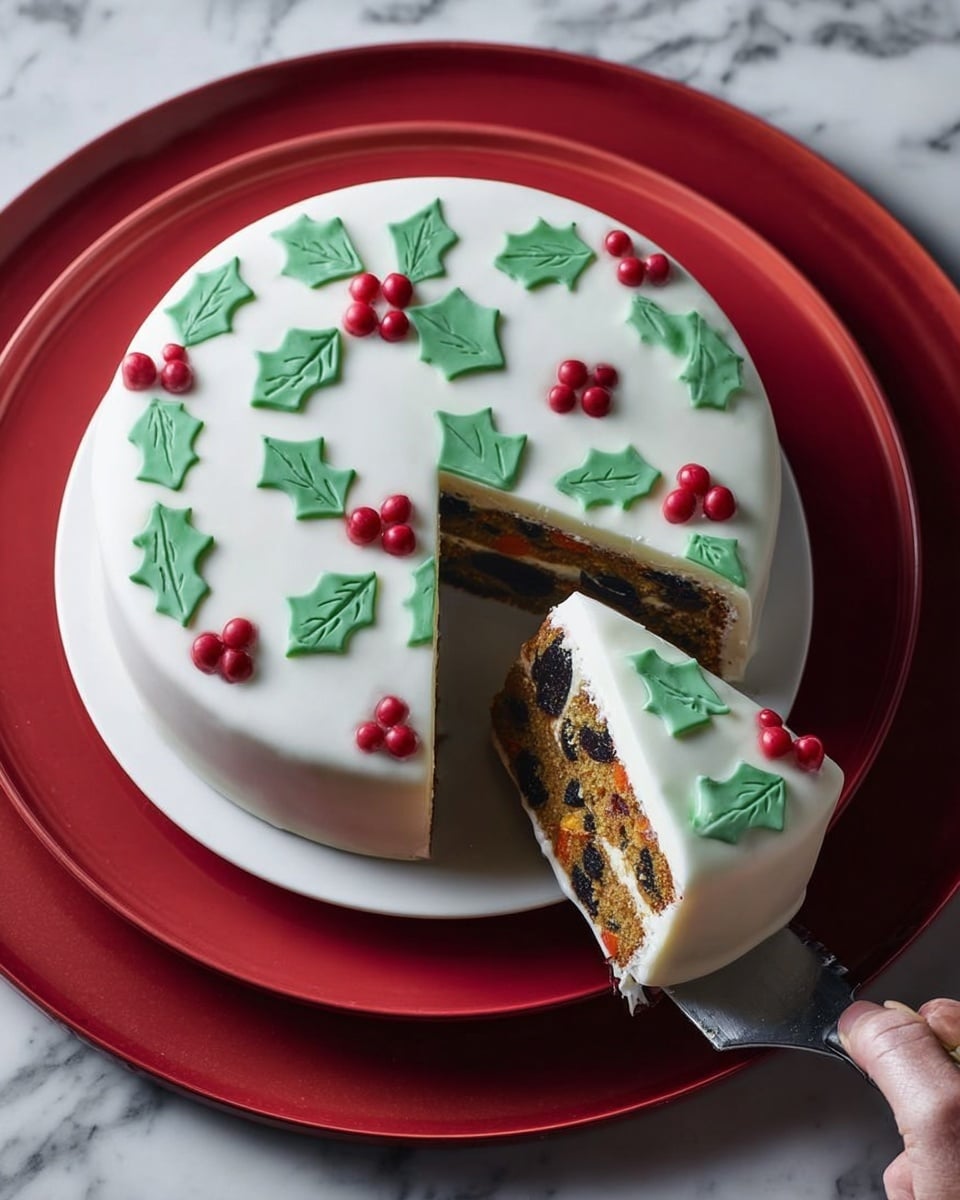 A round cake with a smooth white icing top decorated with flat green holly leaves evenly spread around the surface and small shiny red balls placed between the leaves. One slice has been cut and slightly pulled out, showing multiple layers inside: a golden-brown fruit cake layer filled with dark pieces of dried fruit and nuts, topped by a white icing layer similar to the outside. The cake sits on a white plate placed on a red charger, and the background is a white marbled texture. A woman's hand holds a knife cutting the cake slice. photo taken with an iphone --ar 4:5 --v 7