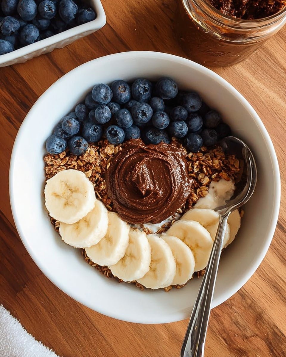 A white bowl filled with a base of crunchy granola forming a ring near the top edge, with a cluster of dark blue blueberries placed in the center. Below the blueberries, a generous dollop of thick, dark brown nut butter sits in the middle. Arranged along the bottom edge inside the bowl, there are evenly sliced, light yellow banana pieces forming a curved line. The bowl is placed on a white marbled surface, with part of a spoon resting on the right side of the bowl. Photo taken with an iphone --ar 4:5 --v 7