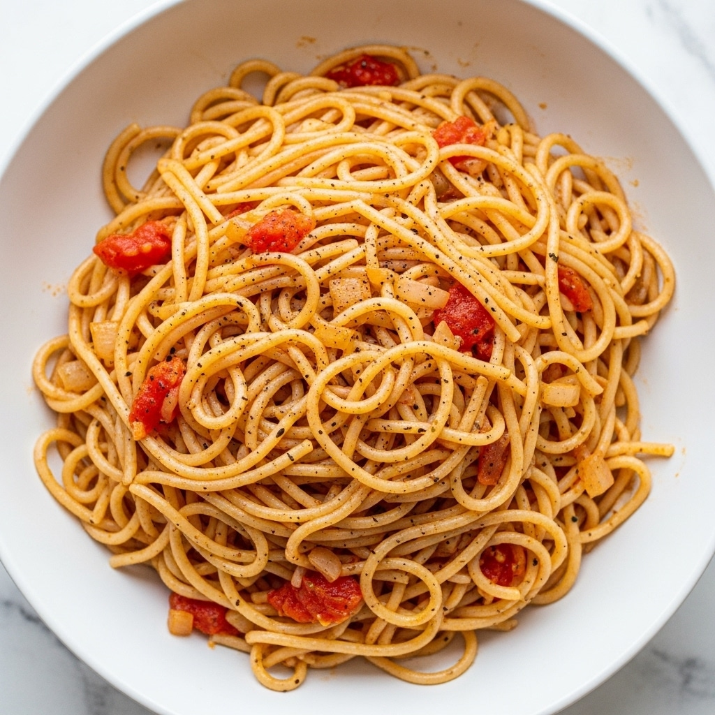 A white plate filled with a generous mound of thin noodles mixed evenly with small pieces of cooked onions and bits of red tomato, giving the noodles light reddish and orange hues. The noodles are coated with spices, giving a slightly speckled texture with darker brown and black specks scattered throughout. The noodles look moist but not wet, layered loosely and piled high in a rounded heap, sitting on a white marbled surface. photo taken with an iphone --ar 4:5 --v 7