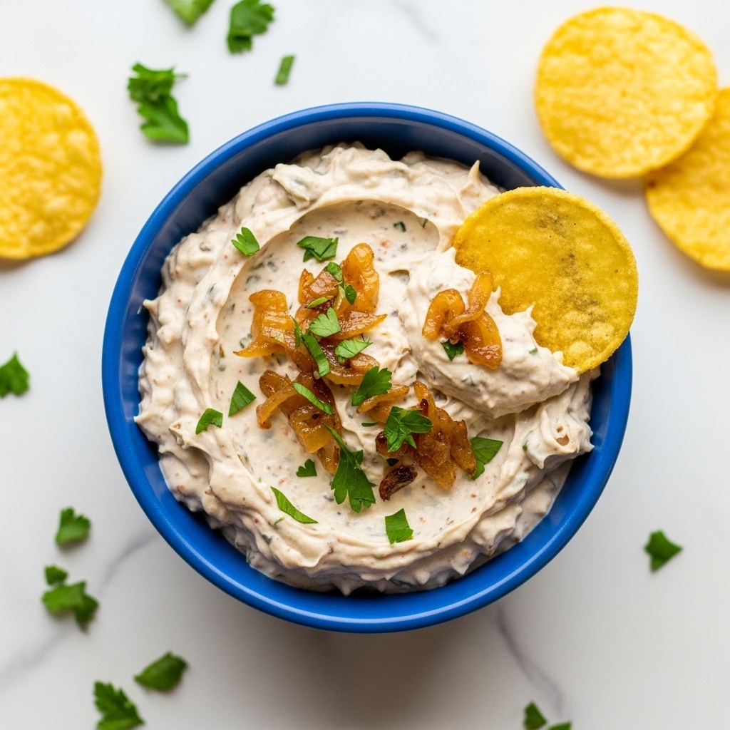 A close-up of a small blue bowl filled with creamy white dip mixed with small browned bits, possibly caramelized onions, and topped with chopped green herbs. The dip has a thick, slightly chunky texture and shows clear swirls where a yellow corn chip is dipped into it, partially covered in the dip. Surrounding the bowl on a white marbled surface are scattered green herb bits and two yellow corn chips, one of which is slightly folded. Photo taken with an iphone --ar 4:5 --v 7