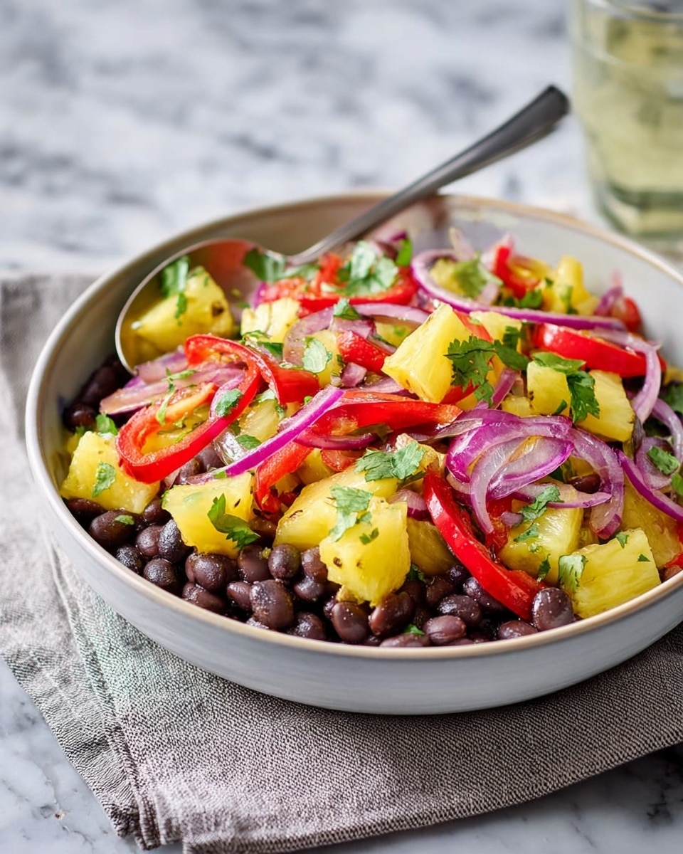 A close-up of a white bowl filled with a colorful salad featuring chunks of yellow pineapple, dark brown beans, sliced red bell peppers, thinly sliced purple onions, and small pieces of green avocado, all mixed with fresh green herbs. The salad rests on a light blue cloth on a white marbled surface, with a spoon partially inside the bowl and blurred glasses in the background. The colors are bright and fresh, creating a vibrant and healthy appearance. Photo taken with an iphone --ar 4:5 --v 7