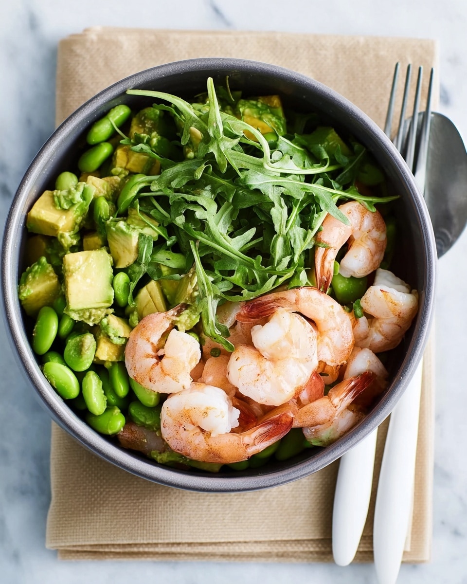 A black bowl filled with a fresh salad sits on a tan napkin over a white marbled surface. The bottom layer of the salad consists of green vegetables like edamame and cut avocado pieces, giving a bright green texture. On top of this, there are several whole cooked shrimp, pale pink in color, placed around the bowl. The shrimp are partially covered with a layer of leafy greens that look fresh and slightly glossy. To the right of the bowl, a white fork and spoon rest on the tan napkin, completing the simple and clean setting. Photo taken with an iphone --ar 4:5 --v 7