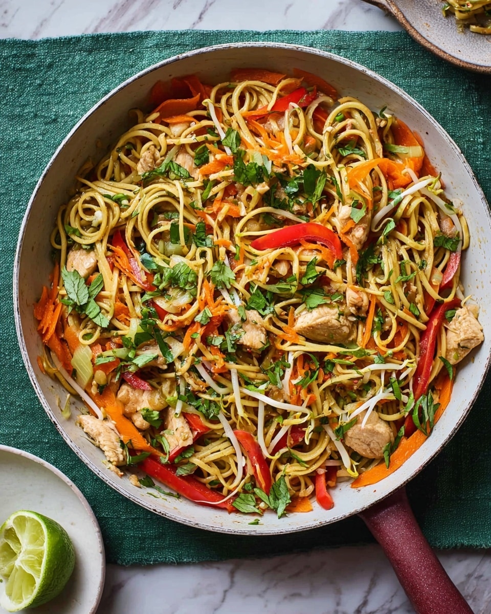 A round white pan filled with a colorful stir-fry showing multiple layers: the bottom layer has light brown noodles mixed with white bean sprouts, the middle layer contains bright orange carrot slices and thin red bell pepper strips, and the top layer is a mix of small pieces of light brown cooked chicken, scattered green herbs, and fresh green basil leaves. The pan has a dark wooden handle and is placed on a pale green cloth on a white marbled surface. To the right of the pan, there is a small white plate with a cut lime on top. photo taken with an iphone --ar 4:5 --v 7