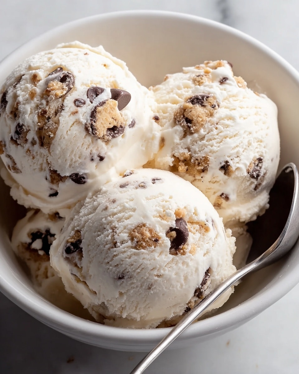 The image shows three large scoops of creamy white ice cream with visible small chocolate chips and chunks of cookie dough mixed evenly throughout. The texture of the ice cream looks smooth and slightly soft, with the cookie pieces adding a rough, crumbly contrast. The scoops are placed close together inside a simple white bowl. A shiny silver spoon is partially visible inside the bowl, resting next to the scoops. The background surface is a white marbled texture. photo taken with an iphone --ar 4:5 --v 7