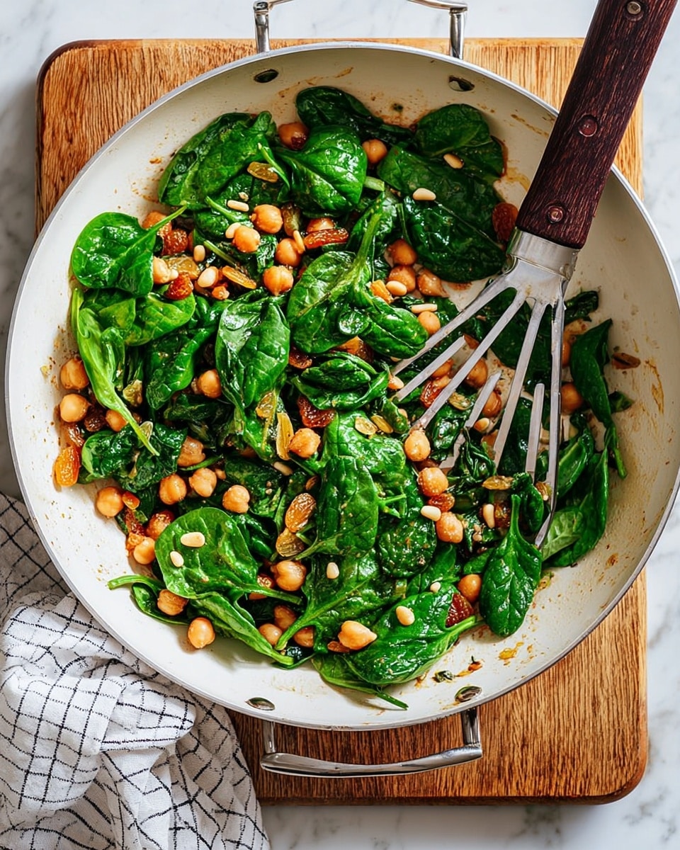 A white pan filled with a colorful mix of cooked chickpeas, bright green spinach leaves, toasted pale yellow pine nuts, and small bits of golden brown caramelized onions. The chickpeas form the base layer, round and orange-tinted from spices, topped unevenly by fresh spinach leaves that are shiny and soft. Scattered pine nuts add small smooth cream-colored highlights on top. A metal slotted spatula rests inside the pan, partially covered by the ingredients. The pan is on a wooden board with a white and gray checkered cloth beneath it, all placed on a white marbled surface. Photo taken with an iphone --ar 4:5 --v 7