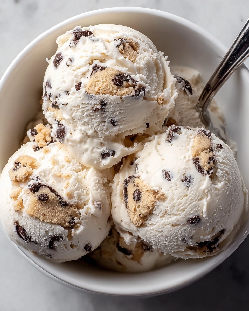 The image shows three round scoops of creamy white ice cream with visible dark chocolate chips and chunks of cookie dough mixed throughout the surface and embedded inside. The ice cream scoops are placed closely together inside a white bowl. A metal spoon is partially inserted into the ice cream on the right side of the bowl. The background is a white marbled texture. The ice cream appears smooth and cold with a slightly soft texture. Photo taken with an iphone --ar 4:5 --v 7