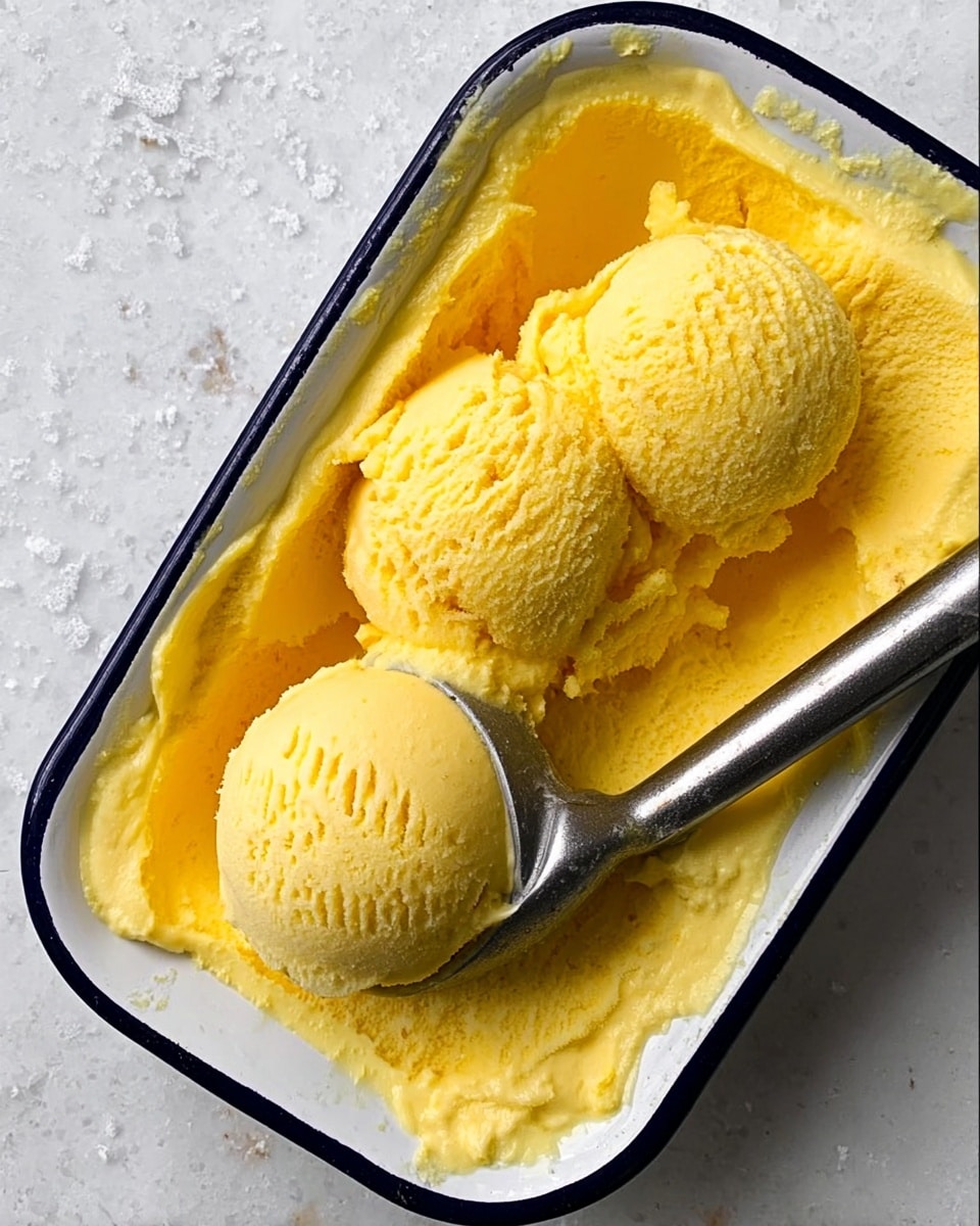 A white enamel rectangular dish with a dark rim holds smooth, bright yellow ice cream, partly melted with three round scoops placed on top. A silver ice cream scoop rests inside the dish, holding one scoop. The texture of the ice cream is creamy and slightly soft. The dish is set on a white marbled surface. Photo taken with an iphone --ar 4:5 --v 7