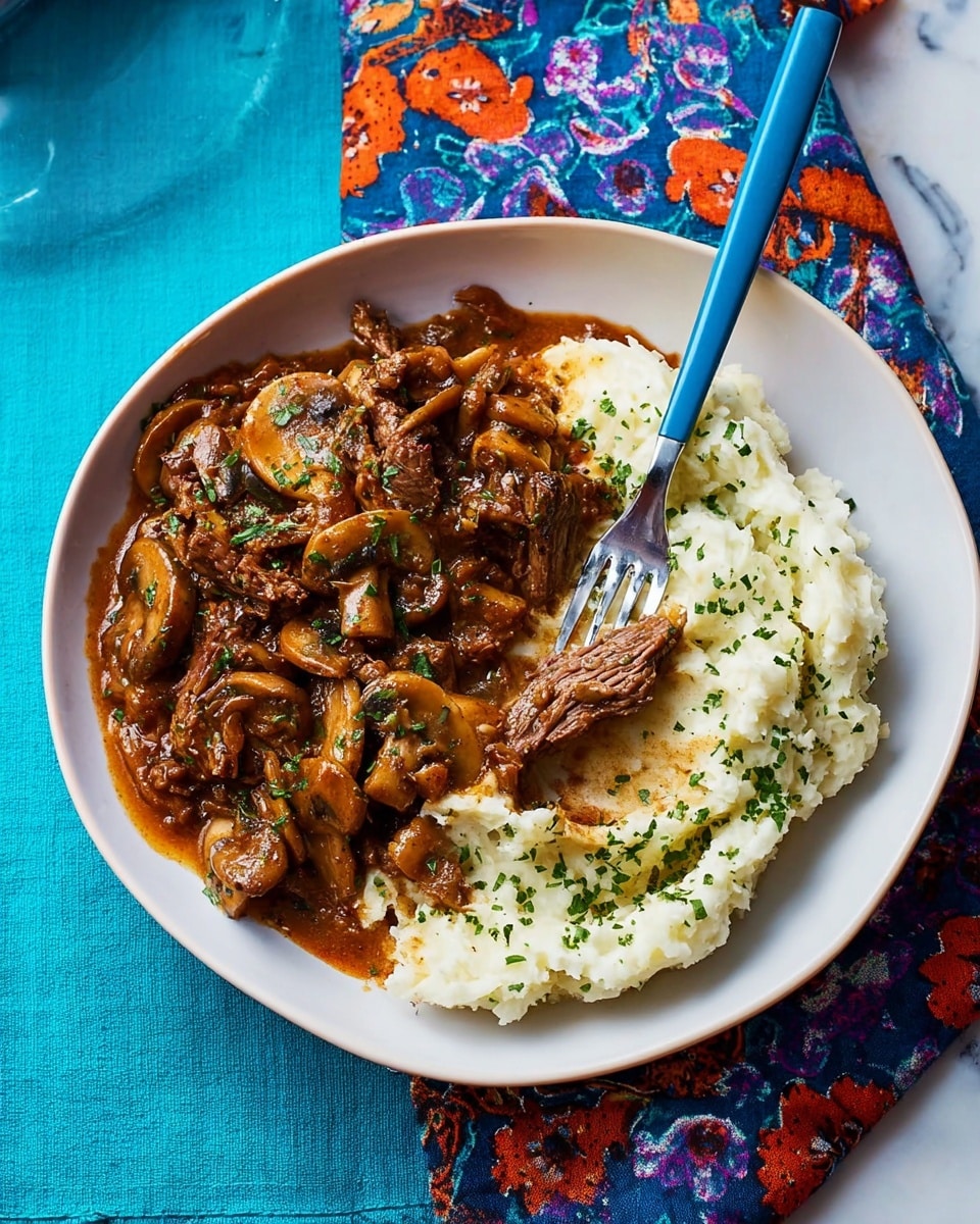 A white plate holds two main layers: on one side, creamy white mashed potatoes sprinkled with green herbs that look like chopped chives and dill, showing a smooth texture with some small lumps; on the other side, a thick brown mushroom and beef stew with visible mushroom slices and tender meat chunks, coated in a rich gravy, slightly overlapping the mashed potatoes. A silver fork with a blue handle rests on the edge of the plate, partly on the stew. The plate is set on a vibrant blue cloth with large flower prints, all placed on a white marbled surface. photo taken with an iphone --ar 4:5 --v 7