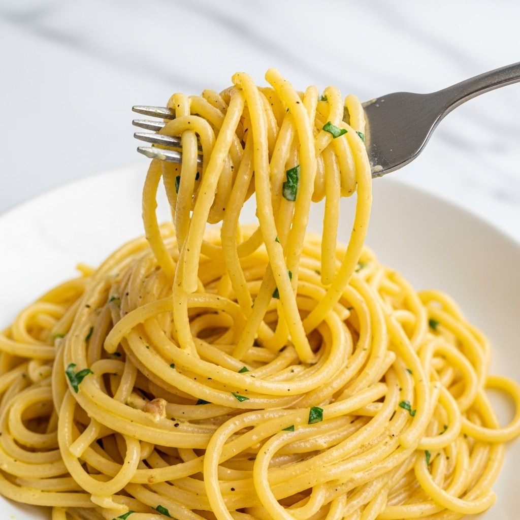 A close-up image shows creamy yellow spaghetti pasta twirled around a silver fork, held by a woman's hand. The pasta strands are smooth and coated with a rich, glossy sauce, speckled lightly with small black pepper bits and green herb pieces. The pasta is piled in layers with some strands loose around, displaying a soft, tender texture. The background is a white marbled texture, making the pasta stand out clearly. Photo taken with an iphone --ar 4:5 --v 7