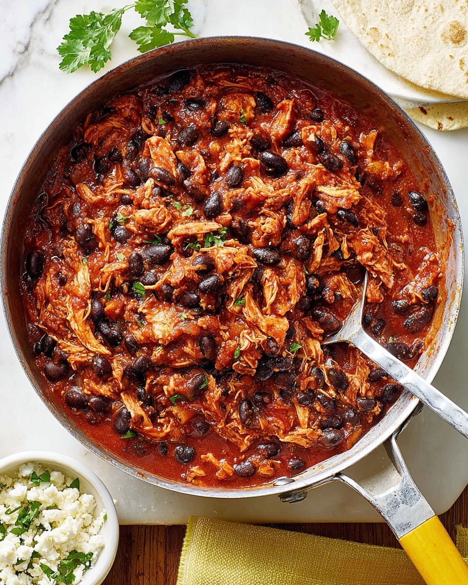 A metal pan filled with a thick stew made of shredded chicken and black beans mixed in a rich, dark red tomato sauce, showing soft textures and small chunks of chicken and beans evenly spread. The pan has a handle with a spoon resting inside it, partially covered by the stew. Next to the pan, there is a partial view of a white bowl with crumbled white cheese and some green herb leaves on the white marbled surface underneath, which is also partly covered by a folded yellow cloth. A tortilla piece is visible in the top right corner. Photo taken with an iphone --ar 4:5 --v 7