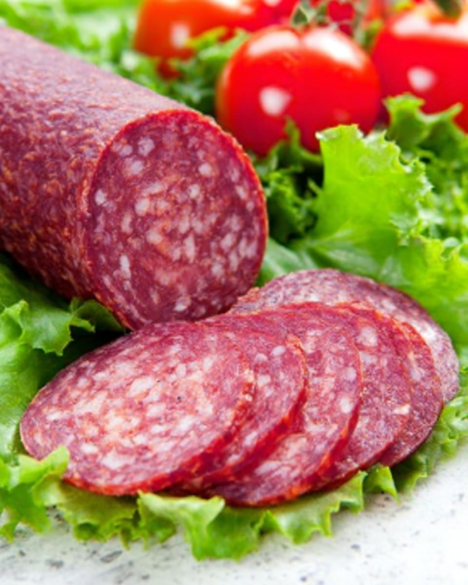 The image shows a close-up view of a sliced sausage placed on a bed of fresh green lettuce leaves. The sausage has a deep red color with small white fat spots inside and a coarse texture on the surface. Five round slices are arranged in a row at the front, each slice showing a marbled mix of red and white. Behind the sausage, there are a few ripe, red cherry tomatoes adding bright color contrast on the green lettuce. All items are set on a clean white marbled surface, creating a fresh and appetizing look. Photo taken with an iphone --ar 4:5 --v 7