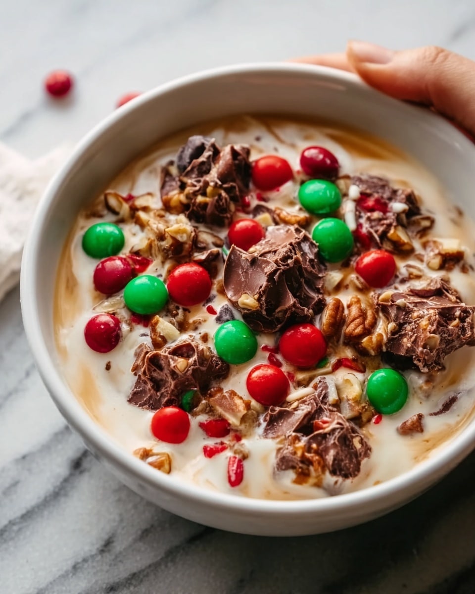 A white bowl filled with a creamy base that looks like vanilla ice cream or yogurt. On top, there are multiple small clusters of dark brown chocolate mousse or fudge creating rough textured piles. Scattered over these are bright red and green candy-coated chocolates adding vibrant color and a smooth, shiny texture. The bowl is placed on a soft fabric, and the background shows a mix of pretzels out of focus. The overall look is a mix of smooth creamy, chunky chocolate, and bright colorful candies. Photo taken with an iphone --ar 4:5 --v 7