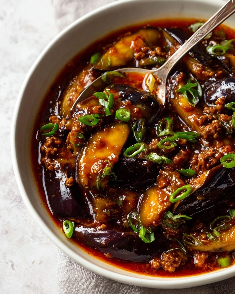 A close-up view of a white bowl filled with a rich, glossy red-brown sauce with tender, cooked eggplant pieces that are dark purple and soft in texture, mixed with small chunks of browned ground meat, and scattered slices of fresh green onion on top, all creating a dense and hearty dish. The spoon inside the bowl has a rustic metallic look and holds a mix of the eggplant and sauce, showing the fullness and thickness of the stew. The bowl is placed on a white marbled surface that contrasts softly with the deep colors of the food. Photo taken with an iphone --ar 4:5 --v 7
