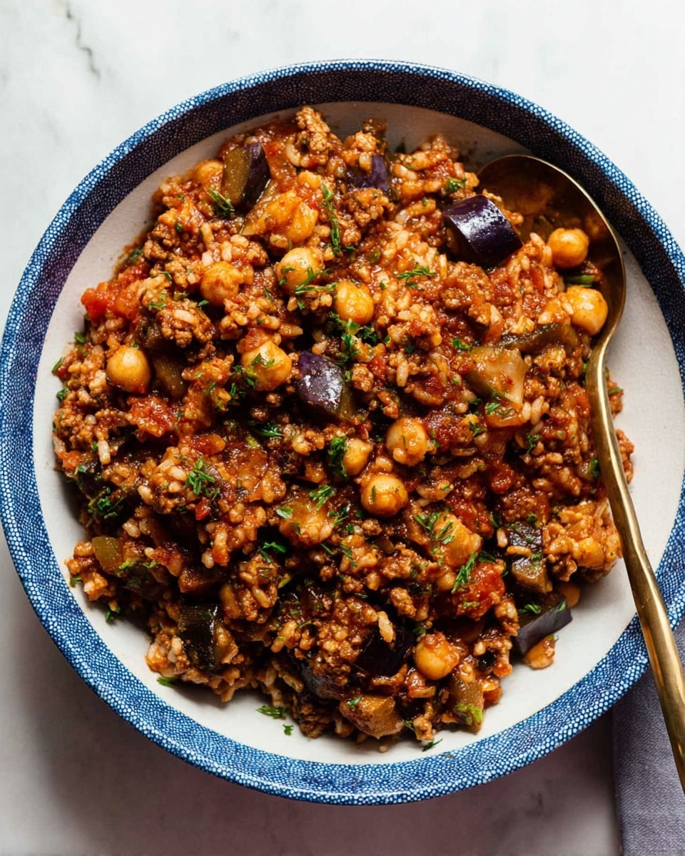 A round white bowl with a blue rim is filled with a mixed dish of cooked rice, small chickpeas, and finely chopped vegetables including eggplant and tomatoes, all coated in a moist, reddish-brown sauce. The dish shows layers of small grains of rice, golden chickpeas, and dark purple eggplant chunks mixed with ground meat and bits of herbs, sprinkled lightly on top. A bronze spoon is placed inside the bowl, leaning on the edge, and the bowl sits on a white marbled surface. Photo taken with an iphone --ar 4:5 --v 7