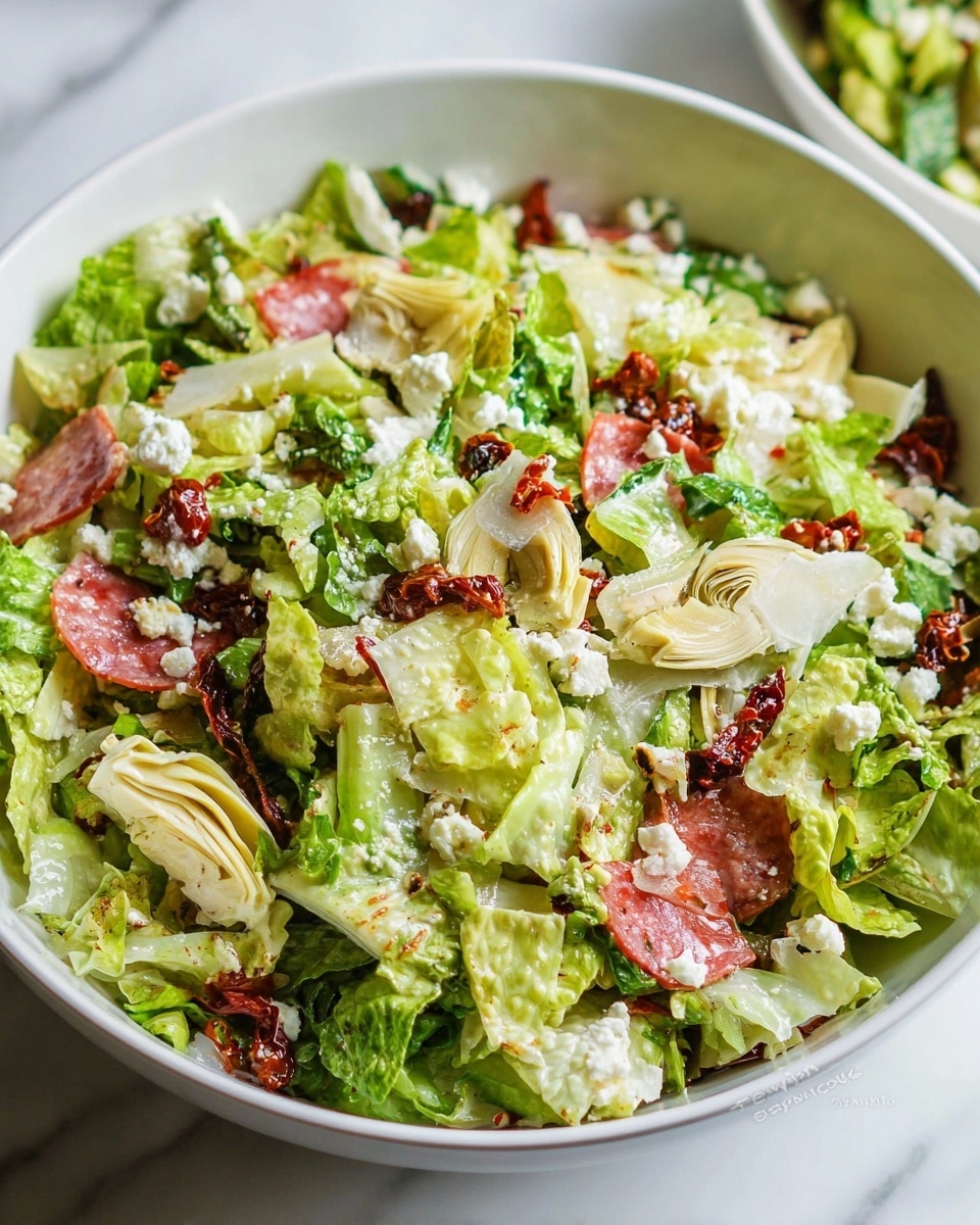 A large white bowl filled with a fresh salad showing three main layers: the base layer of light green chopped lettuce, the middle layer of cream-colored artichoke hearts, and the top layer sprinkled with thin, pink slices of salami, small bits of white cheese, and dark red sun-dried tomatoes. The salad has a mix of rough and smooth textures, with leafy greens, soft artichokes, and thinly sliced meats, all mixed together on a white marbled surface. The background is softly blurred, with a stack of white bowls and a metal citrus juicer visible behind the salad bowl. Photo taken with an iphone --ar 4:5 --v 7