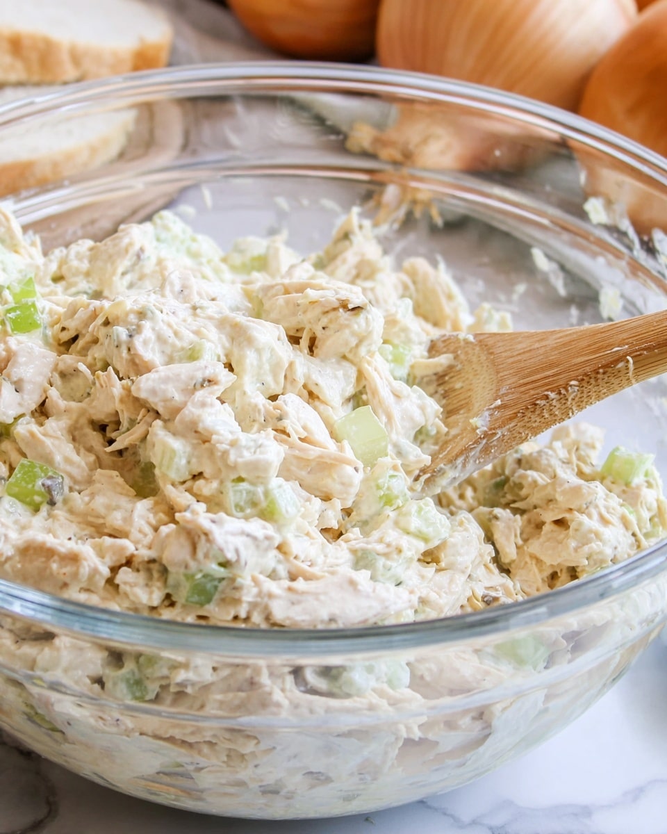 A clear glass bowl filled with a creamy chicken salad mixture, showing roughly shredded white and light brown chicken pieces coated in a white dressing. Light green celery chunks are mixed evenly throughout, adding small pops of color and a crunchy texture. A wooden spoon is partially submerged on the left side, lifting some of the salad. In the blurred background, a whole onion and a loaf of bread are visible, with everything set against a white marbled surface. photo taken with an iphone --ar 4:5 --v 7