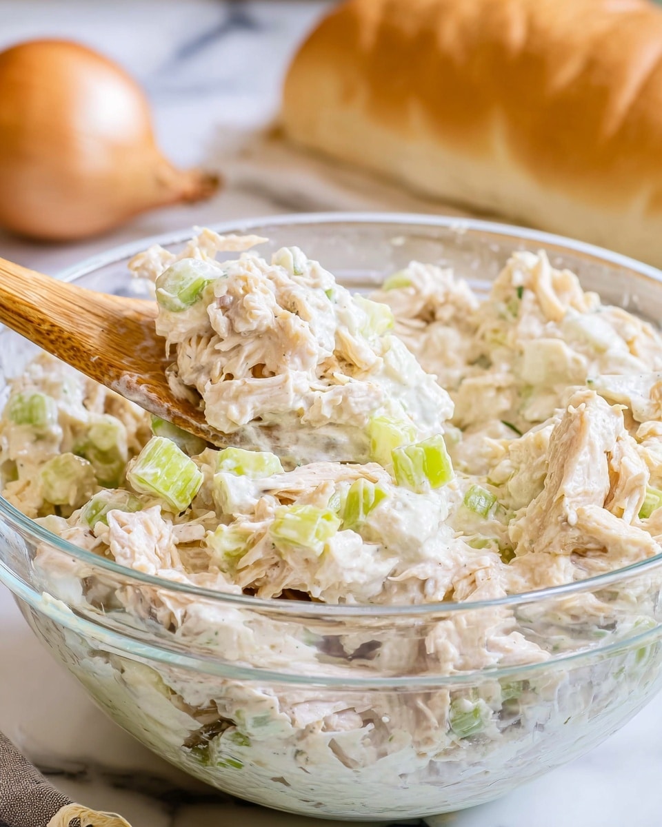 A clear glass bowl filled with a creamy, chunky chicken salad mixed with small pieces of light green celery and pale yellow onions, showing a mix of white and beige textures, with a wooden spoon partially submerged into the salad. The bowl sits on a white marbled surface, and in the background, blurry onions and bread are visible. Photo taken with an iphone --ar 4:5 --v 7