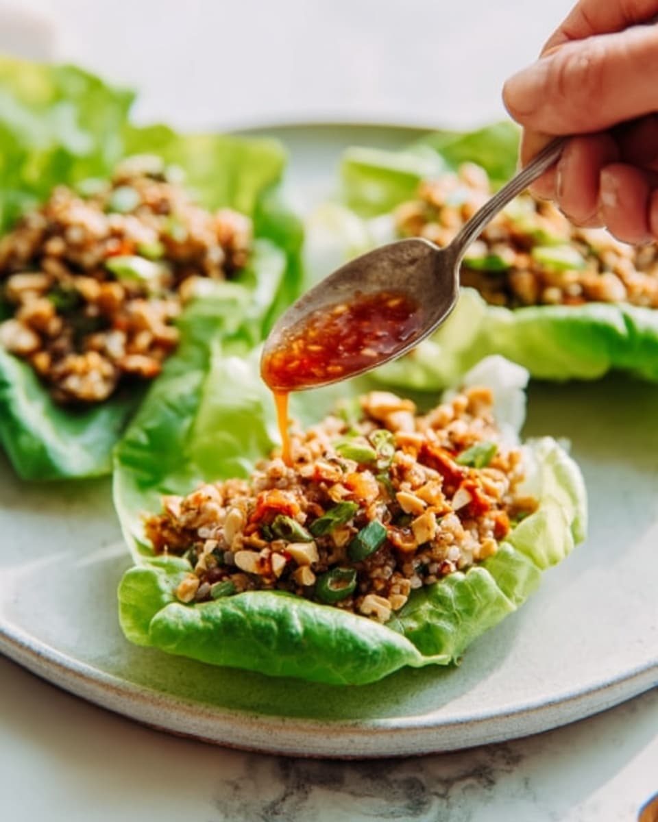 A white plate with two fresh green lettuce leaves placed side by side, each leaf holding a mix of finely chopped ingredients that look like cooked chicken or tofu, herbs, and nuts. The top layer of the mixture is sprinkled with crushed peanuts. A woman's hand is holding a spoon above the left lettuce leaf, pouring a reddish sauce over the filling. The background is a white marbled surface. Photo taken with an iphone --ar 4:5 --v 7