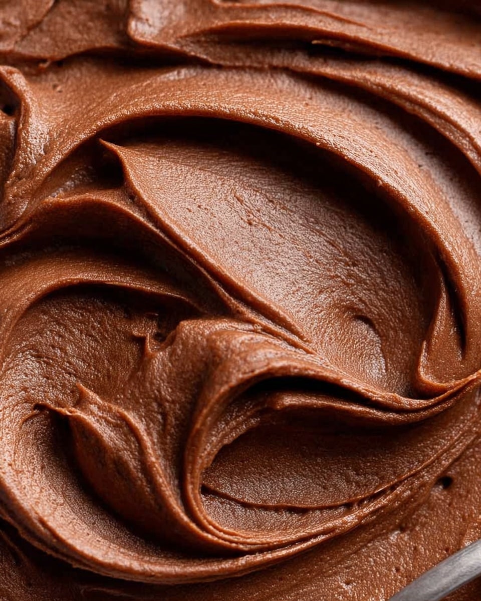 A close-up image shows a creamy, smooth layer of rich chocolate frosting, thickly spread with soft swirls and gentle peaks creating a velvety texture. The frosting is dark brown with glossy highlights, filling most of the view. A small part of a silver spoon is visible at the bottom edge, touching the frosting which looks soft and easy to spread. The background has a white marbled texture. Photo taken with an iphone --ar 4:5 --v 7