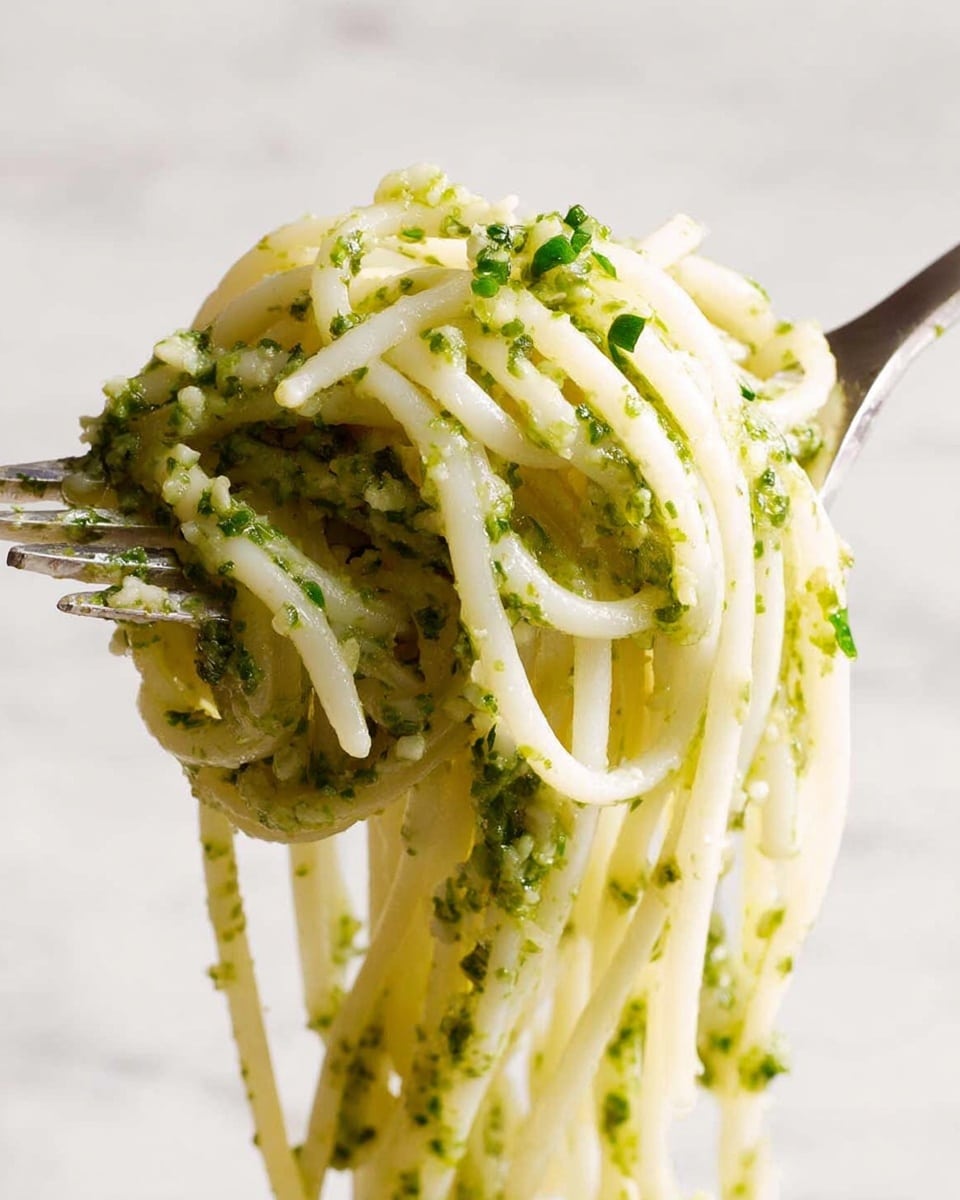 A close-up of thin white noodles coated with a green pesto sauce held in metal tongs, showing long strands of pasta dripping with thick, chunky green sauce made of herbs and small bits, all set against a white marbled texture background. photo taken with an iphone --ar 4:5 --v 7
