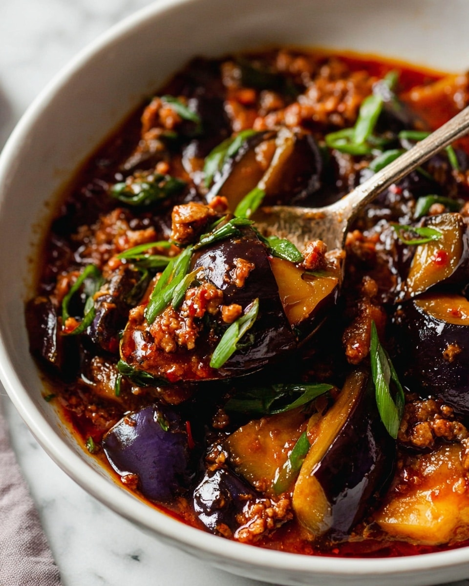 In a white bowl filled with rich, dark reddish-brown sauce, there are large, glossy pieces of eggplant coated in the sauce, mixed with small bits of browned ground meat. The dish is topped with thinly sliced green onions scattered evenly over the glossy sauce surface. A tarnished silver spoon rests inside the bowl, lifting a mix of eggplant and sauce, hinting at the thick, sticky texture of the dish. The bowl sits on a white marbled textured surface with soft natural lighting. photo taken with an iphone --ar 4:5 --v 7