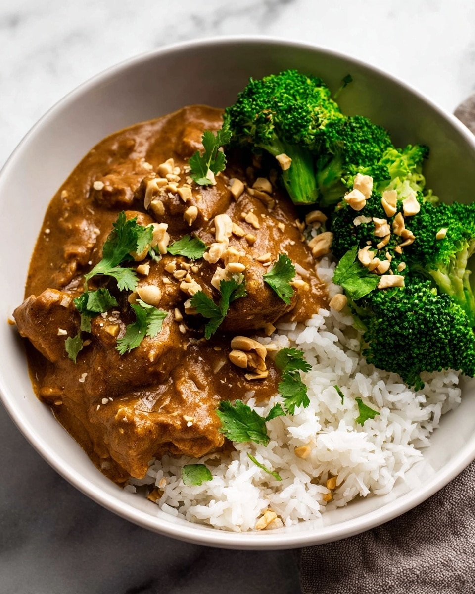 This image shows a white bowl filled with three layers of food. The bottom layer is white rice, fluffy and spread evenly across the bowl's base. The middle layer is a creamy brown curry sauce with chunks of tender meat, covering most of the rice and topped with some chopped cilantro and crushed nuts. The top layer is bright green steamed broccoli florets seasoned with seeds, arranged on one side of the bowl. The bowl is placed on a soft brown cloth over a white marbled surface. Photo taken with an iphone --ar 4:5 --v 7