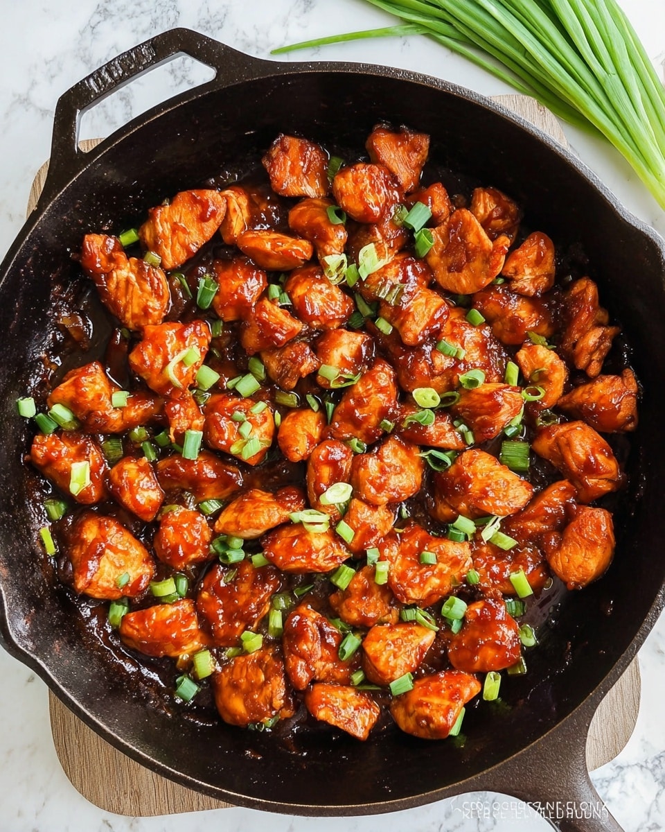 A black cast iron skillet filled with cooked bite-sized pieces of chicken, each piece coated in a shiny, reddish-orange sauce. The chicken pieces are scattered evenly across the skillet, with small bits of sauce pooled around them. On top of the chicken, there are small, bright green chopped scallions adding a fresh color contrast. The skillet rests on a white marbled surface with some green onion stalks partially visible on the side. The texture of the chicken appears soft and tender with a slight glaze from the sauce. photo taken with an iphone --ar 4:5 --v 7