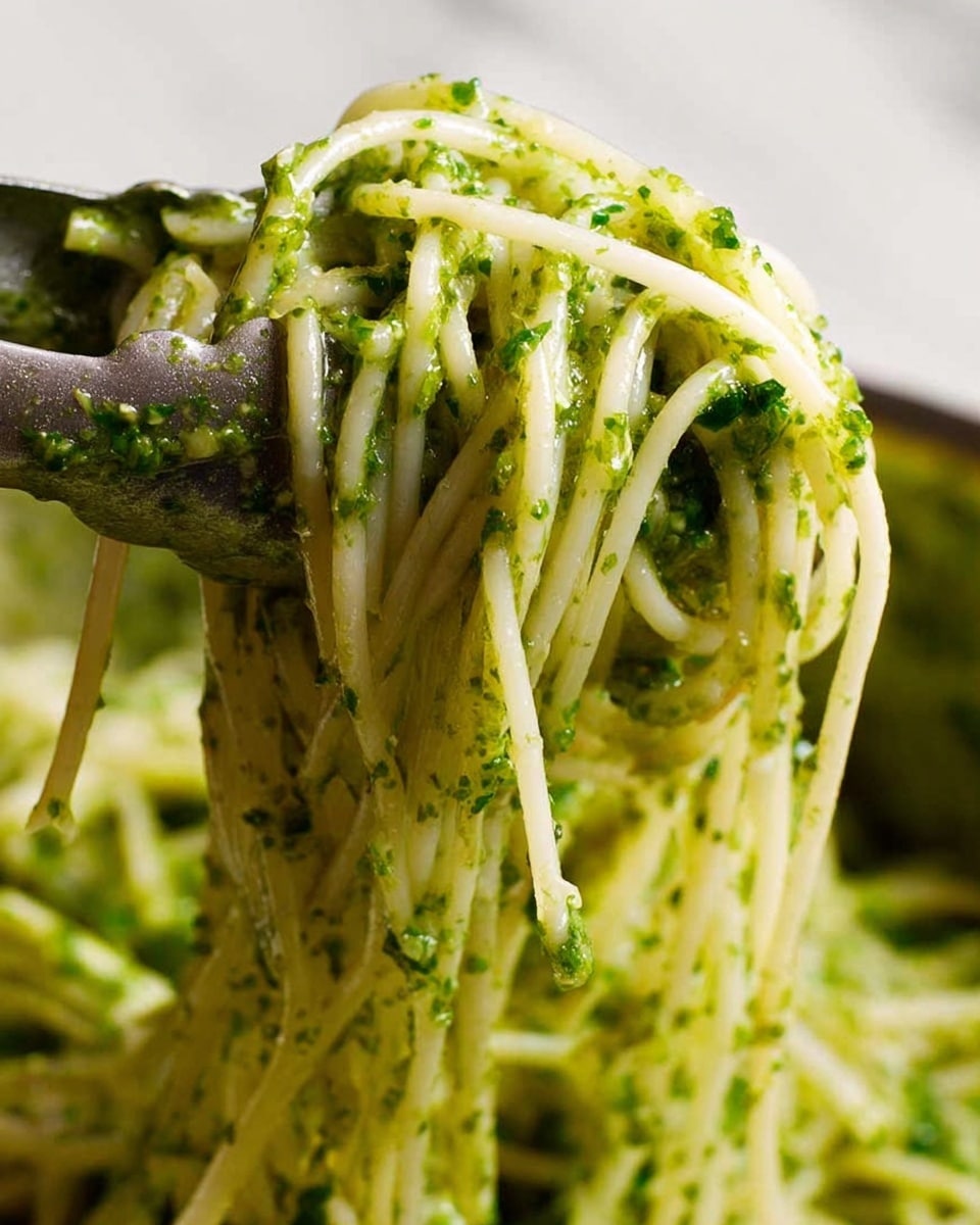 A close-up view shows a cluster of thin white noodles wrapped around a fork, covered in small patches of bright green pesto sauce with visible herb bits. The noodles are glossy and smooth, hanging loosely with a slight shine, while the sauce offers a textured contrast with specks of herbs. The background features a clean white marbled texture, making the pasta and sauce colors stand out clearly. Photo taken with an iphone --ar 4:5 --v 7